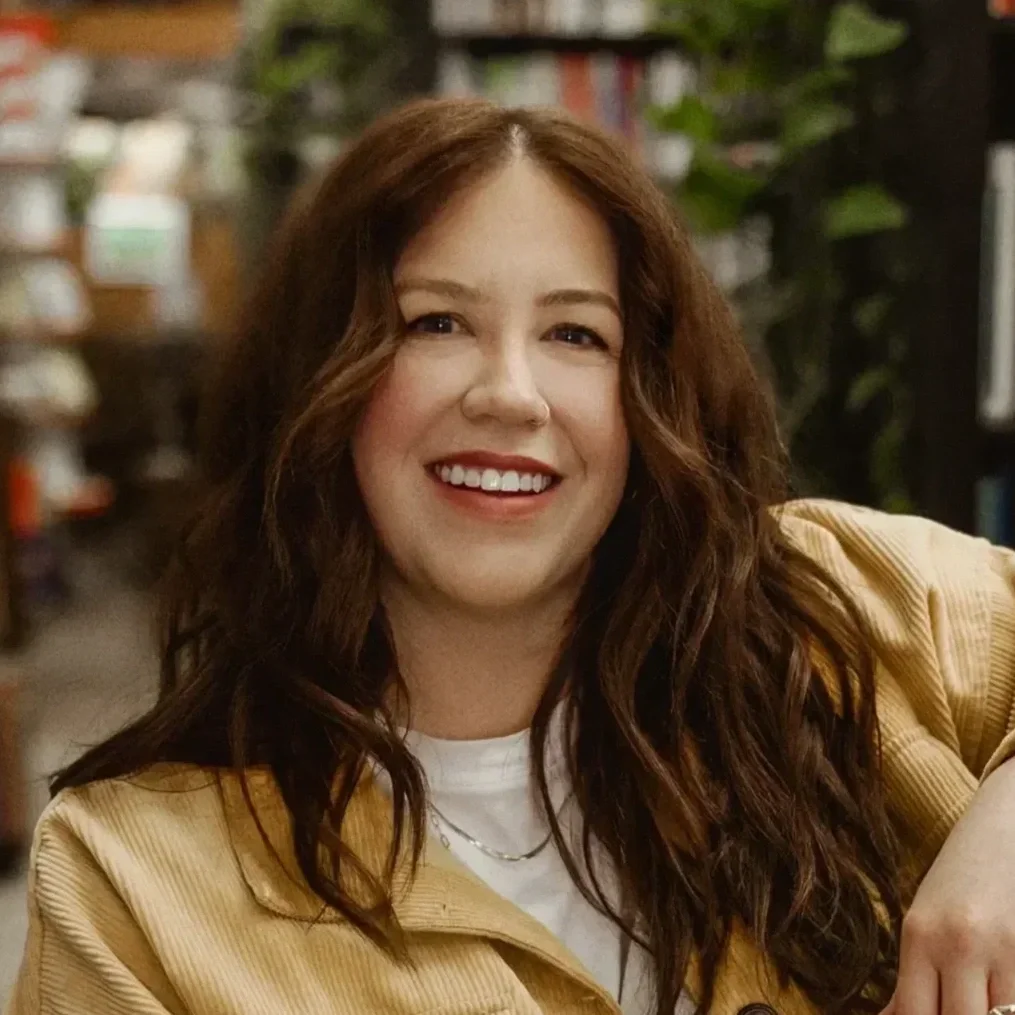 A woman with long, wavy brown hair and a bright smile, wearing a yellow jacket and a white shirt, in a bookstore or library setting.