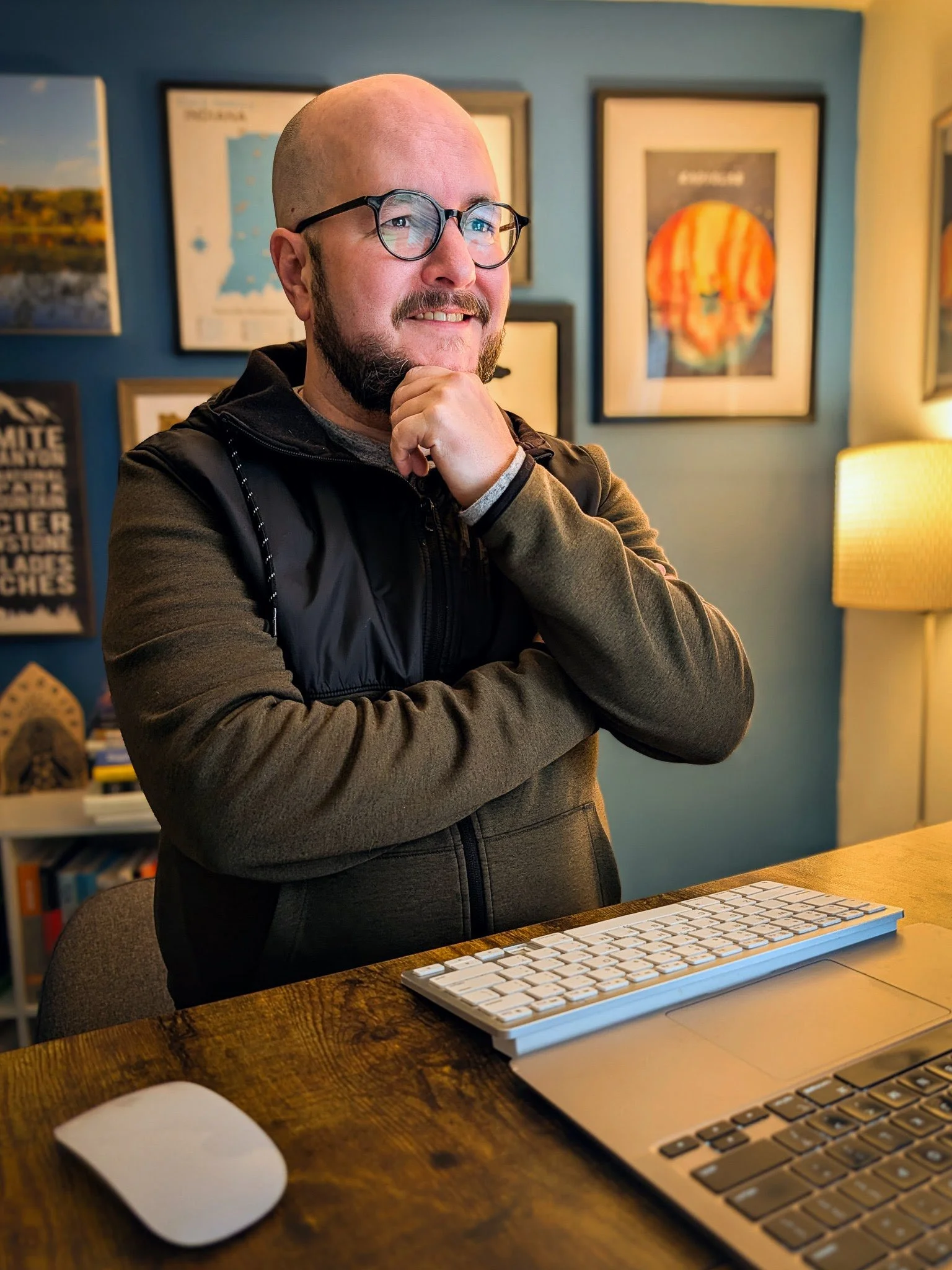 Grief coach, Adam Bouse, standing behind a desk with a keyboard and a mouse, in a room decorated with framed pictures on a blue wall.