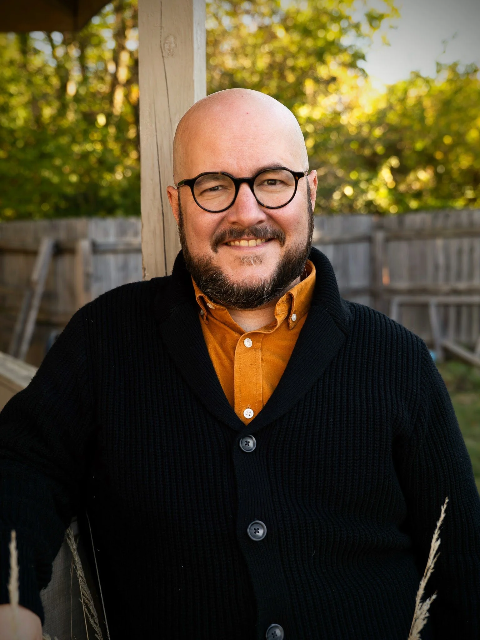 Professional headshot of Adam Bouse, grief coach, wearing an yellow button-up shirt and a black sweater, standing outdoors near a wooden post with a fence and trees with green leaves in the background.