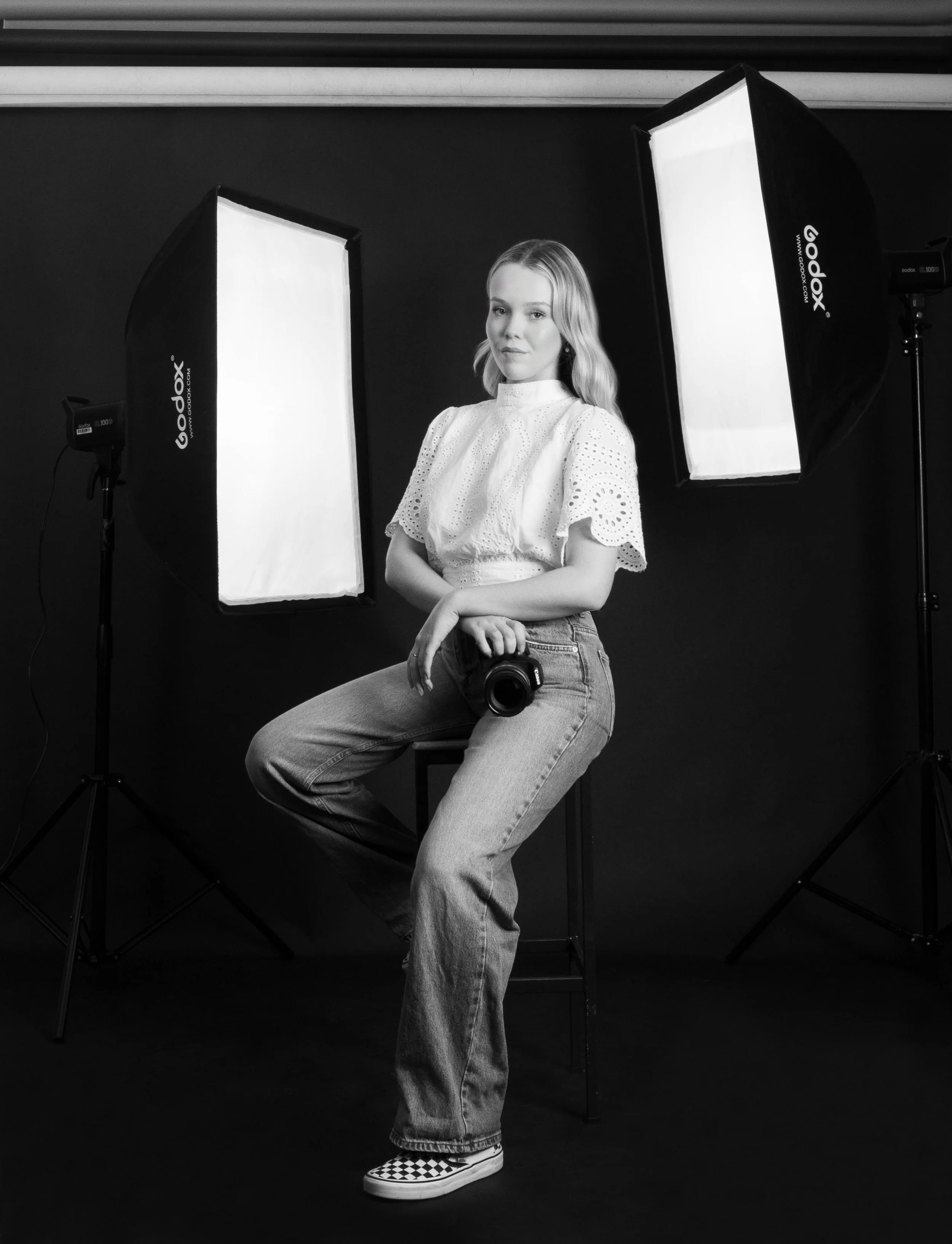 Black and white photo of a young woman with wavy blonde hair, wearing a white eyelet blouse and jeans, sitting on a stool in a photography studio with two large softbox lights and a dark background. She is holding a camera in her lap and looking at the camera.