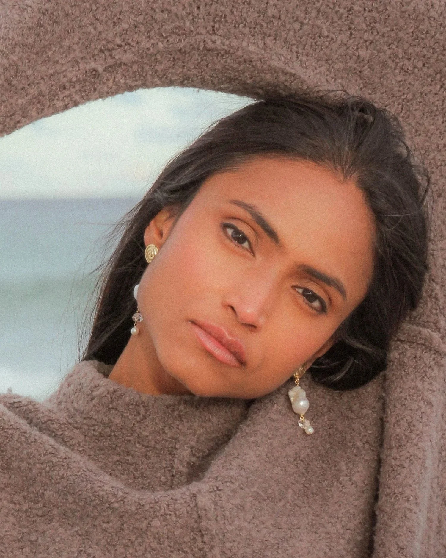 A woman with dark hair styled in loose waves, wearing gold earrings and a pearl drop earring, dressed in a brown fuzzy sweater, lying on a textured surface with a blurred background.