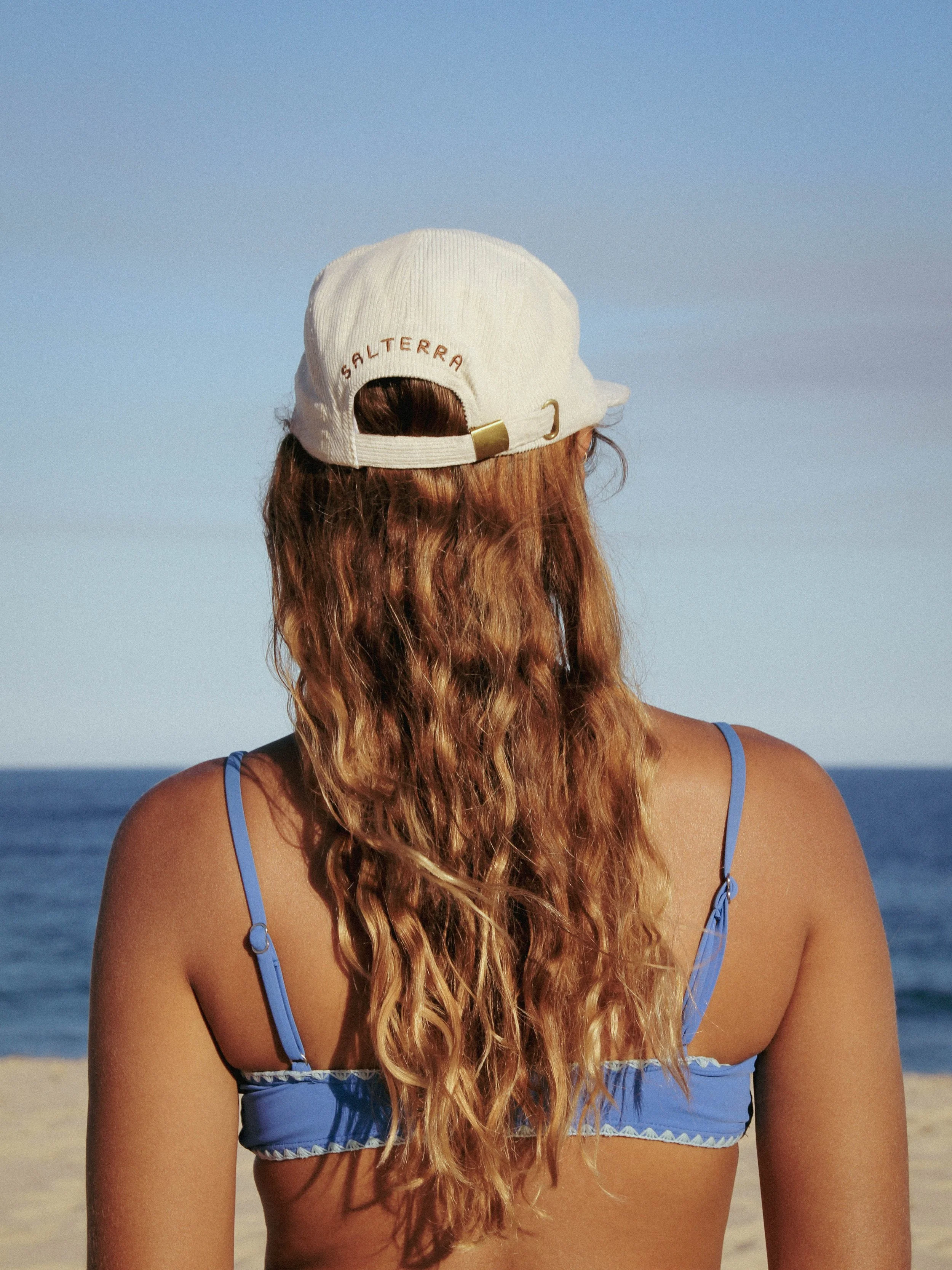 A woman with long, wavy red hair stands on a beach, facing away from the camera, wearing a white baseball cap with the word 'Saltera' embroidered on the back and a blue bikini top. The ocean and sky are visible in the background.