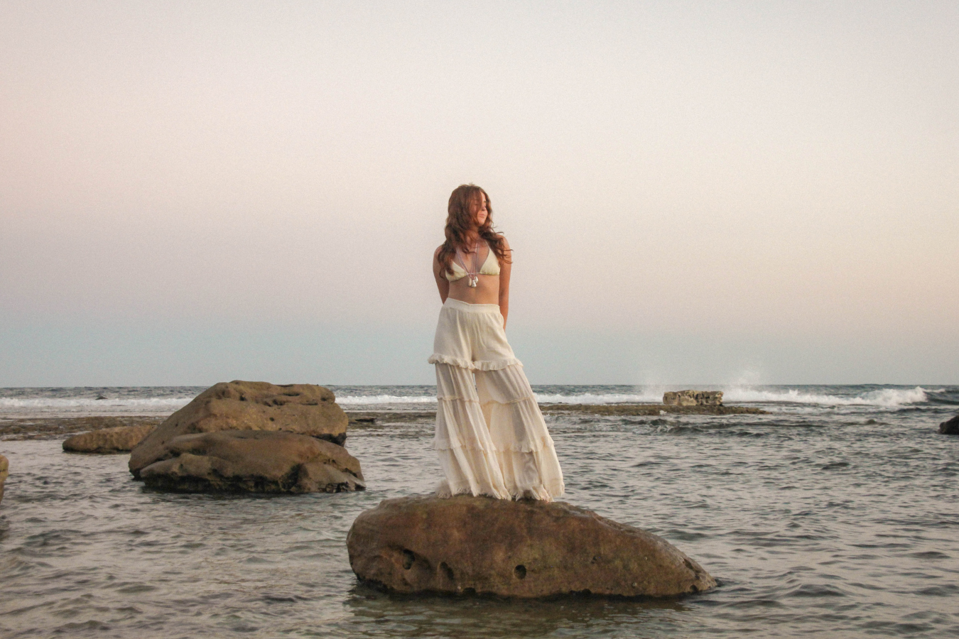 A woman in a light yellow bikini top and long white skirt stands on a rock by the ocean at sunset, with waves and rocks visible in the background.