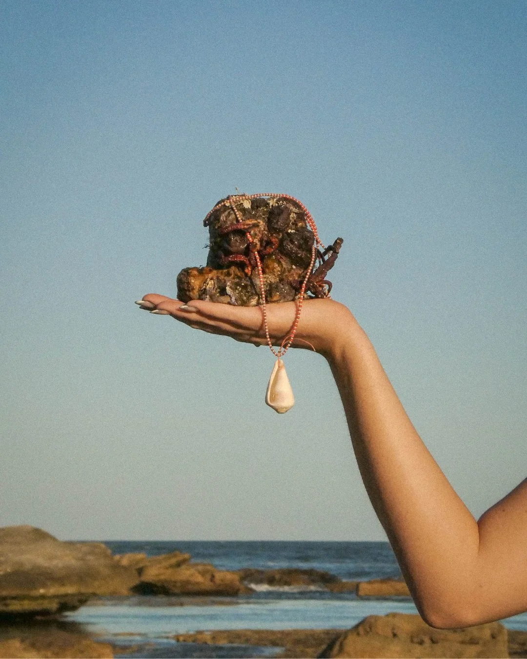 A person holding a decorated stone or sculpture with beads and a pendant, outside by the ocean.