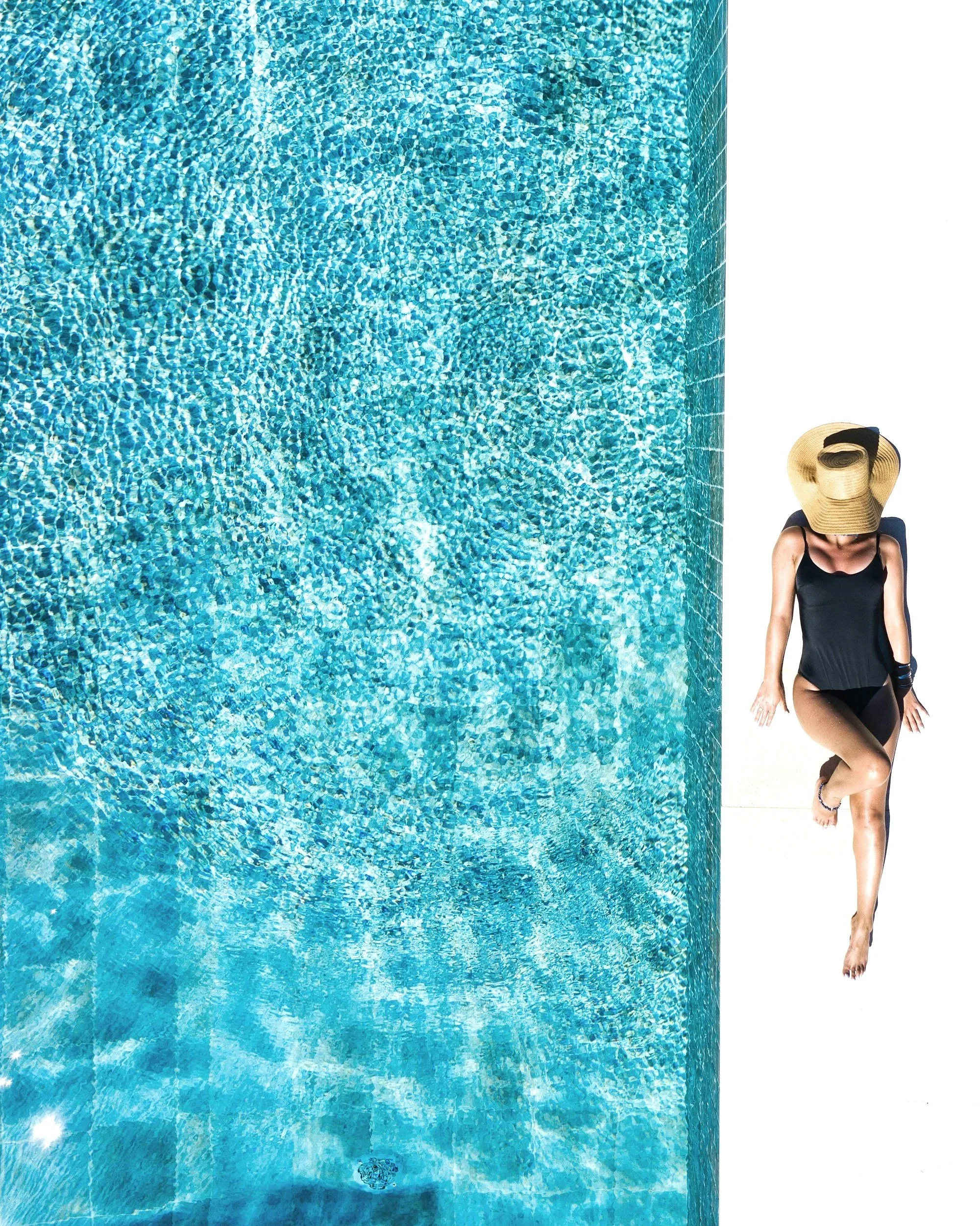 Woman wearing a black swimsuit and straw hat, sitting on a white surface with her head bowed down, against a plain white background.