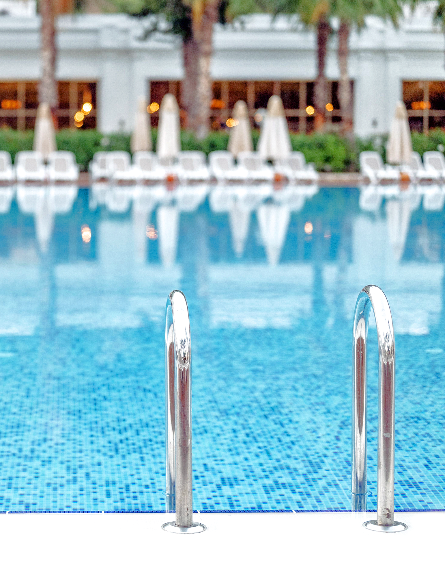 Empty swimming pool with metal ladder at the foreground, surrounded by numerous white lounge chairs and umbrellas, with a building and trees in the background.