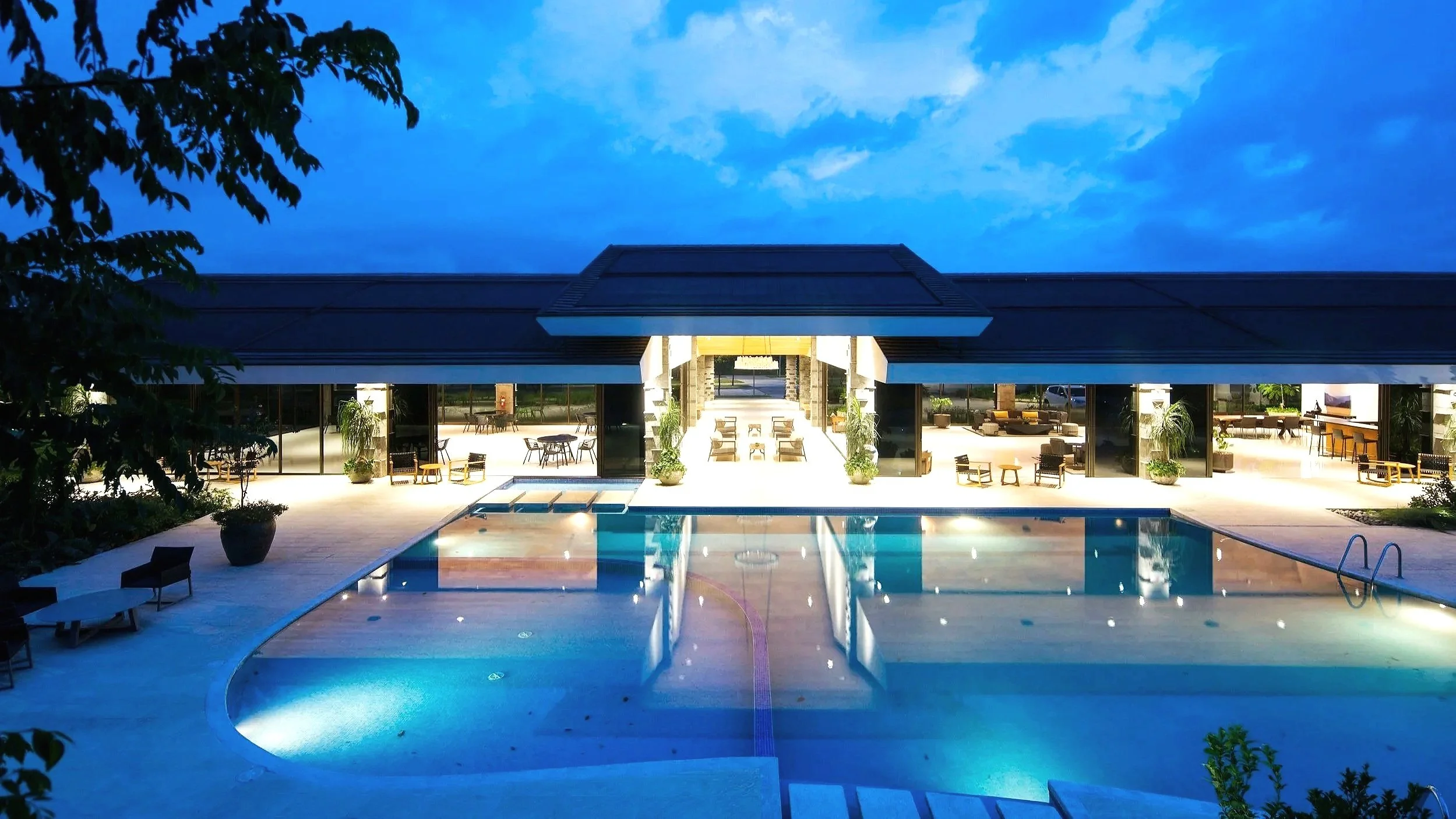 A modern house with large glass windows and doors, outdoor seating area with chairs and tables, a pool with built-in lights, and landscaping with potted plants, all illuminated during dusk.