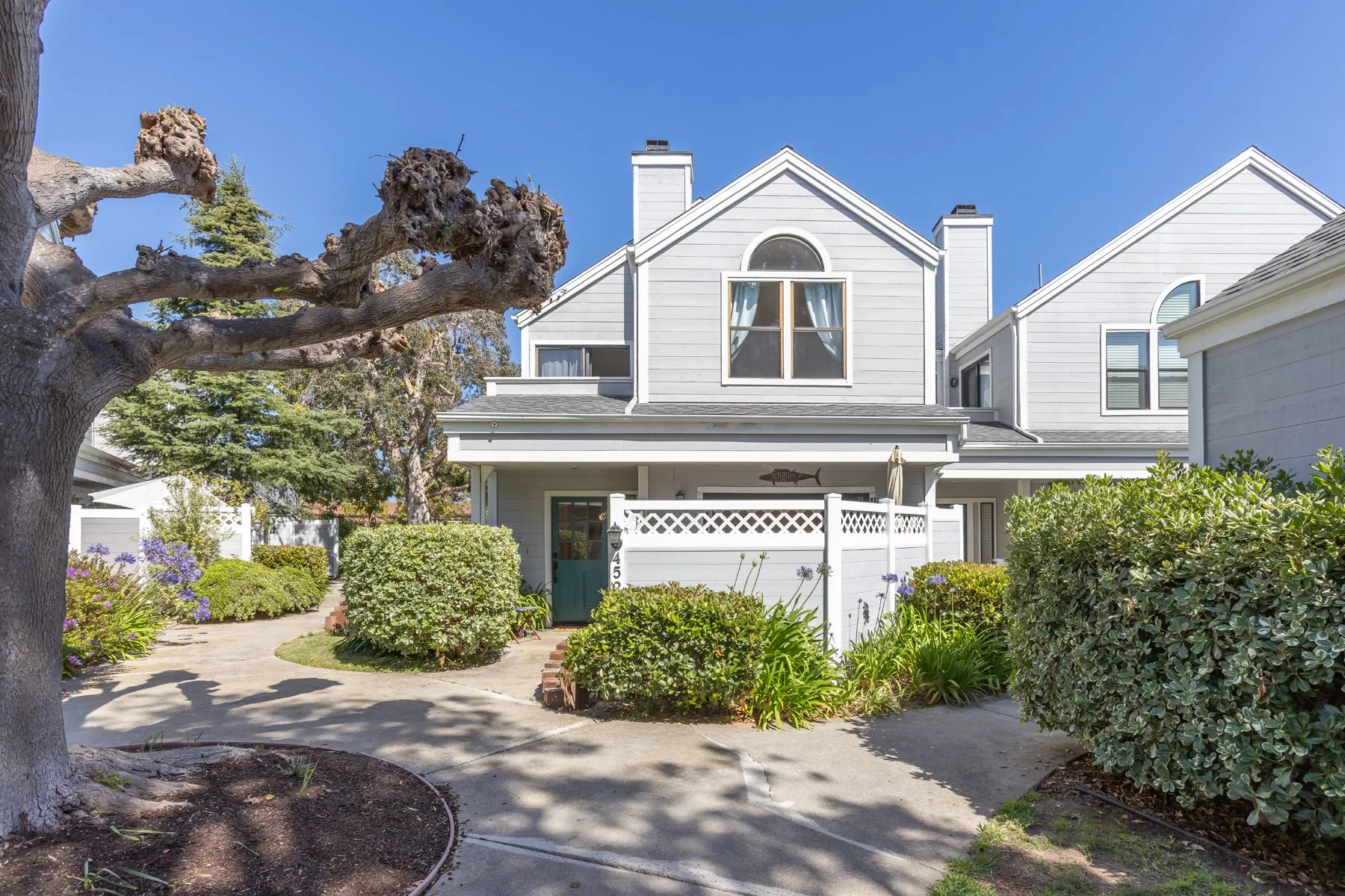 A white two-story house with a front yard filled with green bushes, purple flowers, and trees. There is a sidewalk leading to the front door, which is painted teal. The house has a gabled roof, a large window above the porch, and a white wooden fence. The sky is clear and blue.