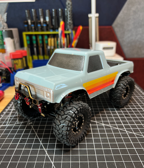 Remote control off-road RC truck with large tires and colorful stripes on the body, placed on a grid-pattern work surface in a workshop setting.
