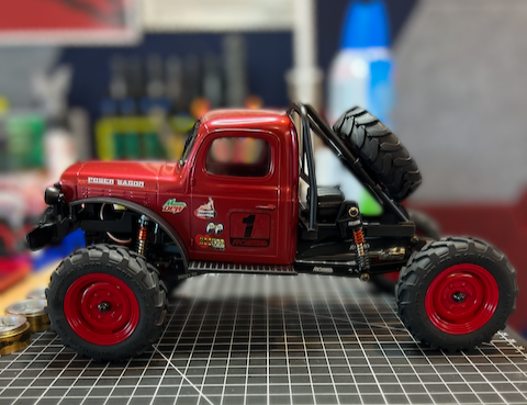 Red remote-controlled off-road RC truck with large tires and a spare tire mounted on the back, on a workbench with tools in the background.