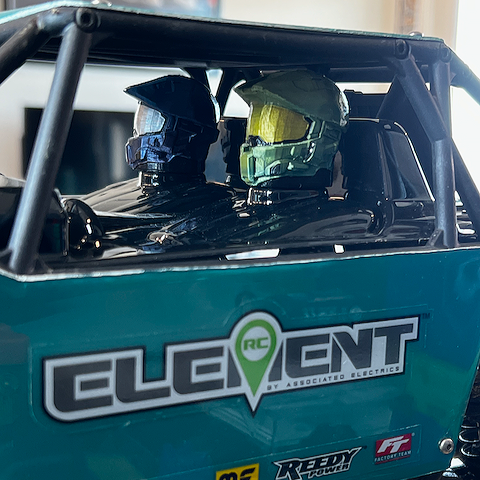 Two helmets, in the style of Master Chief from Halo, one blue and one yellow, inside a teal RC off-road vehicle with logos on the side.