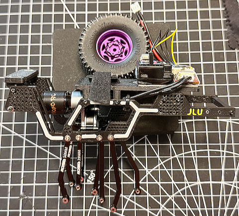 A remote-controlled car chassis with a visible motor, electronic components, and a purple wheel, placed on a black grid cutting mat.