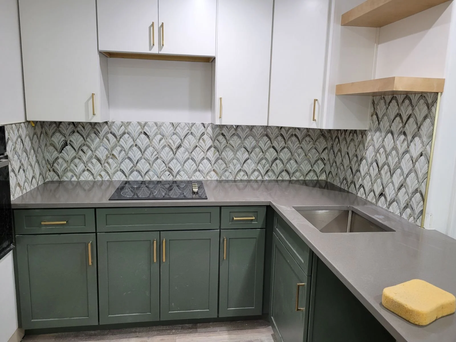 Kitchen with green lower cabinets, white upper cabinets, patterned backsplash, gray countertop, built-in stove, and sink, with a yellow cleaning sponge on the counter.