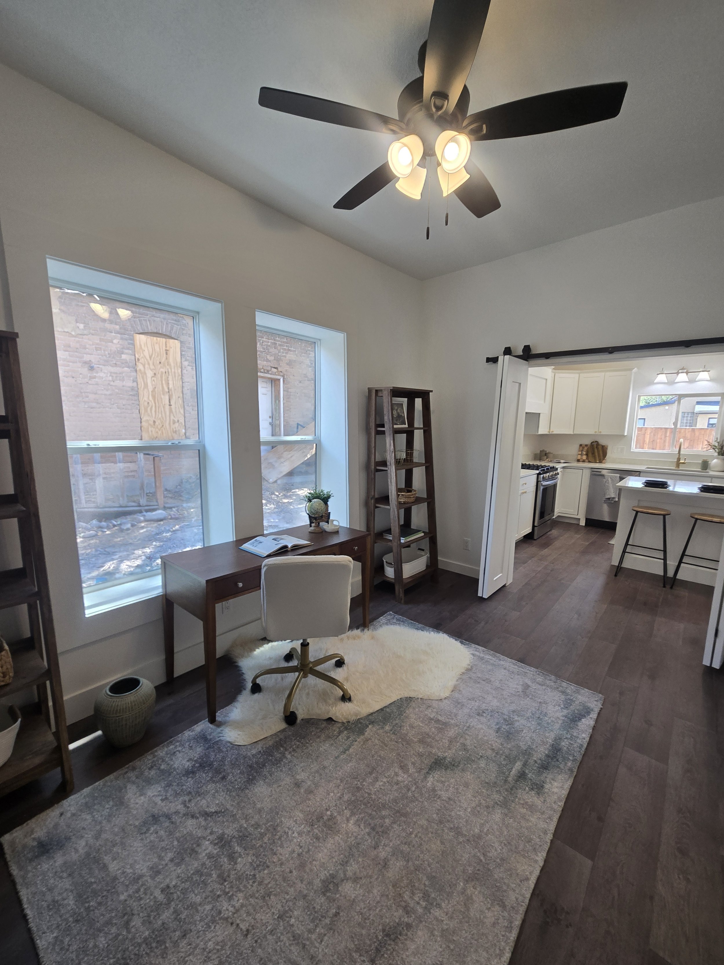 Living room with large windows, white walls, a white fluffy rug, small desk with a chair, a wooden bookshelf, and a ceiling fan with lights. The kitchen is visible in the background.