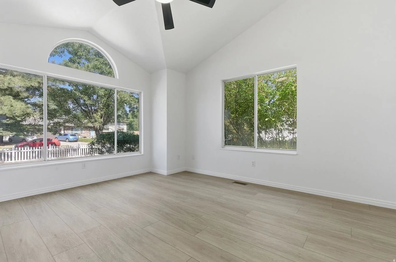 Empty room with large windows, white walls, light wood flooring, and a black ceiling fan.