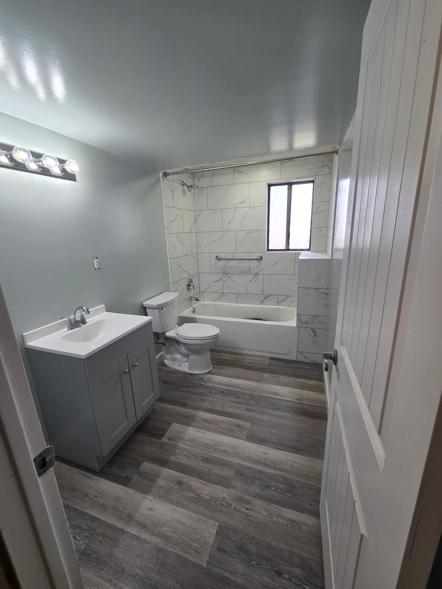 Bathroom with a gray vanity, white sink, toilet, bathtub with marble tiles, window, and wood-look flooring.
