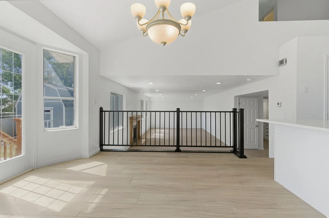Empty bright living room with light-colored wooden floors, large windows, a fireplace, and a black safety gate separating an upper level from the lower level. A chandelier hangs from the ceiling.