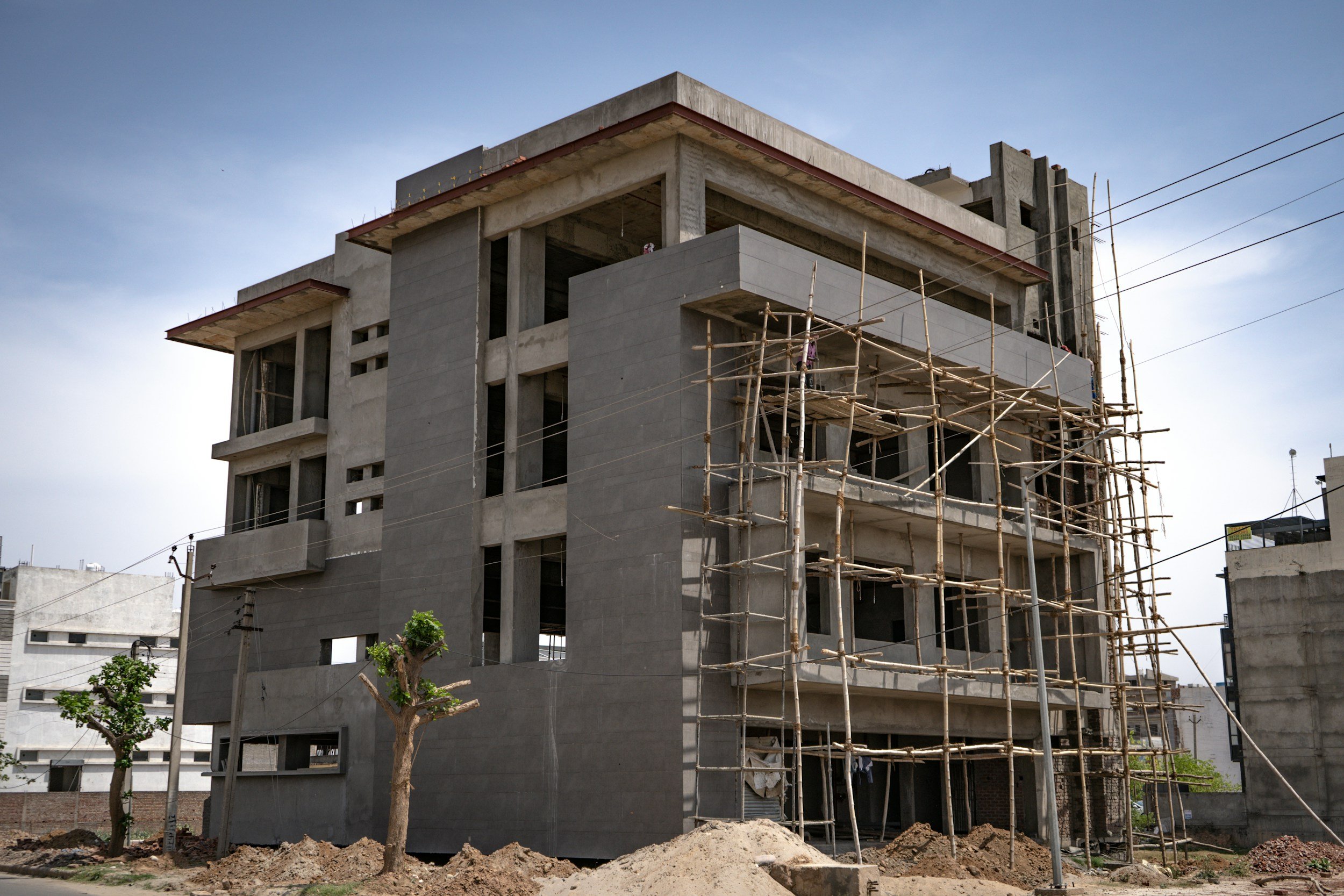 A multi-story building under construction with scaffolding on the front and multiple window openings. The building's exterior is unfinished, exposing concrete structure, with a few small trees and mounds of dirt at the base, set against a clear blue sky.