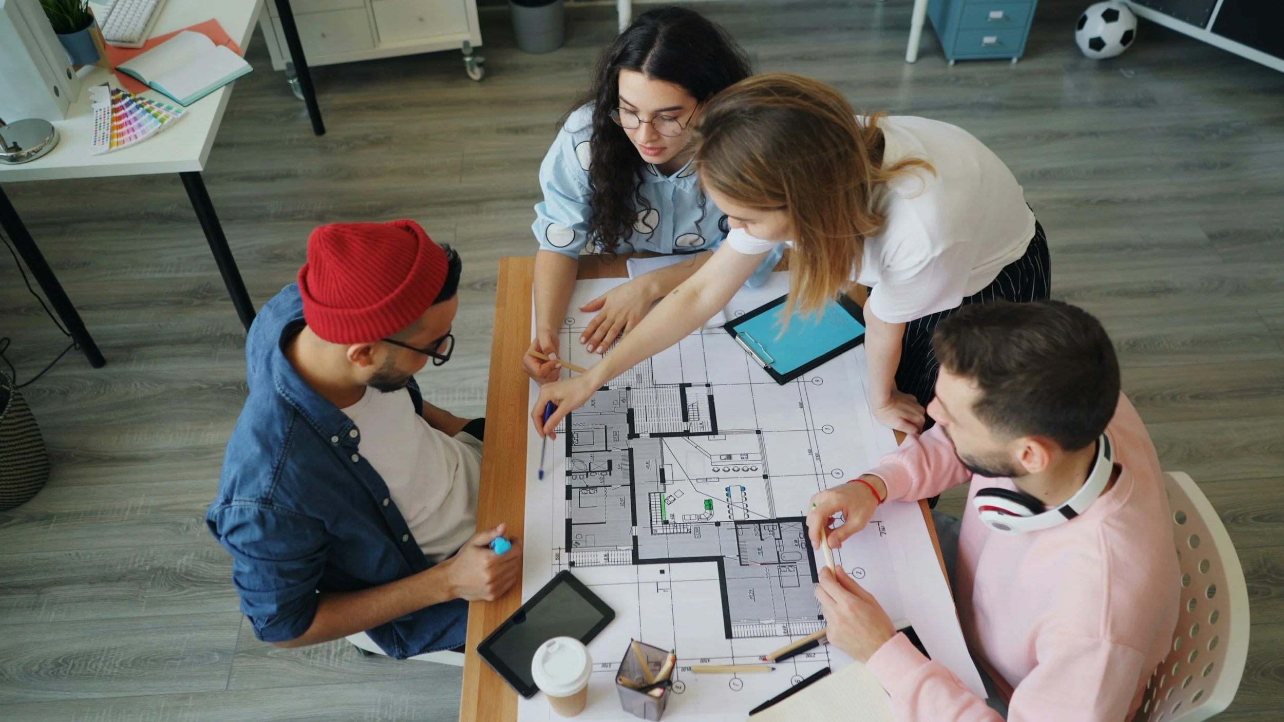 Four people gathered around a table reviewing an architectural floor plan. They are focused on the blueprint, with one pointing at it. The table has tablets, a smartphone, coffee, and stationery. The setting appears to be a modern office or workspace with a bookshelf and decorative items in the background.