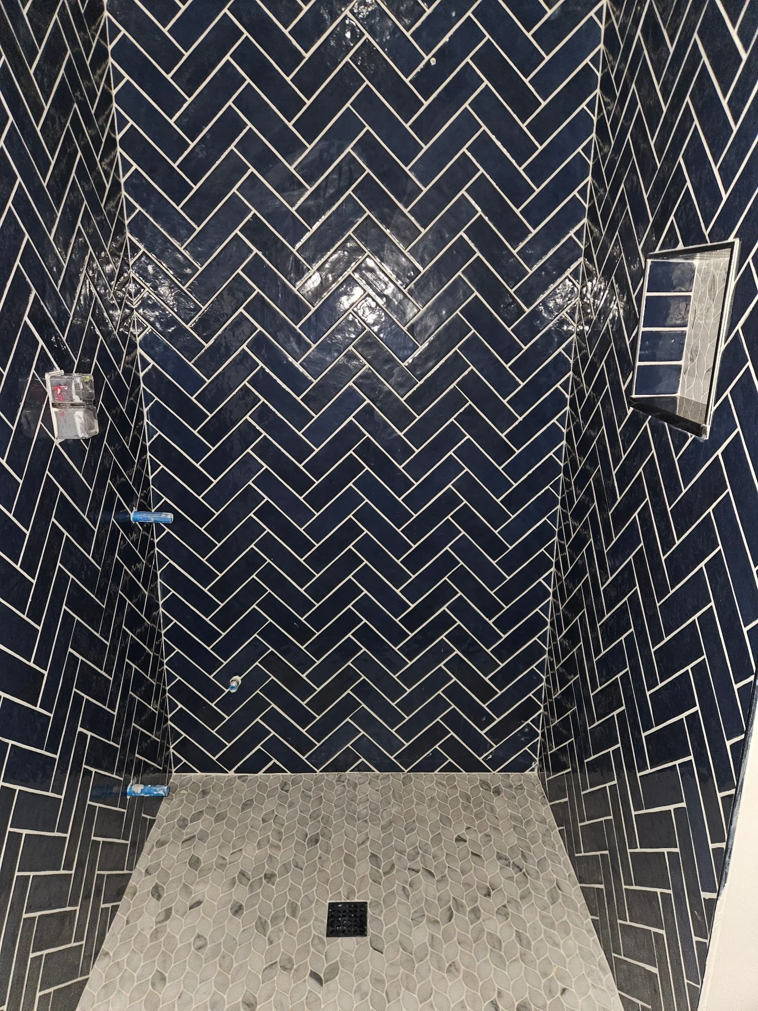 Shower area with dark herringbone tiles on the walls and light patterned tiles on the floor, with a small black drain in the center.