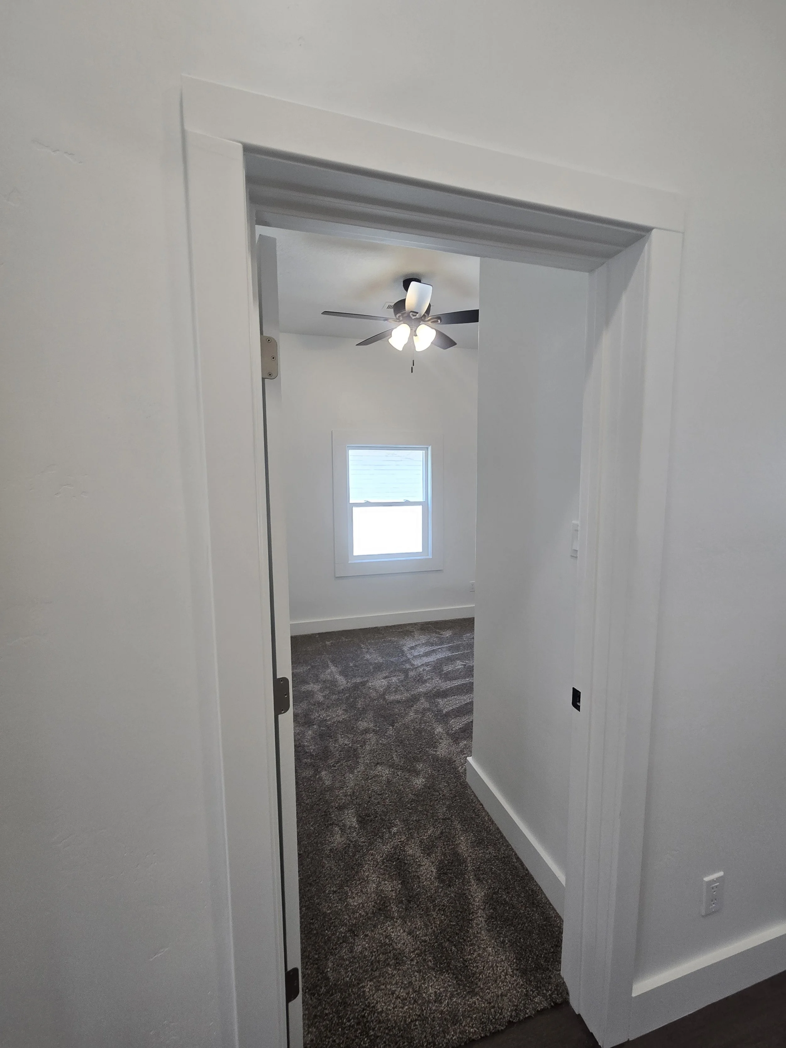 View through a doorway into a small room with gray carpet, a white ceiling with a ceiling fan and light, a window with closed blinds, and plain white walls.