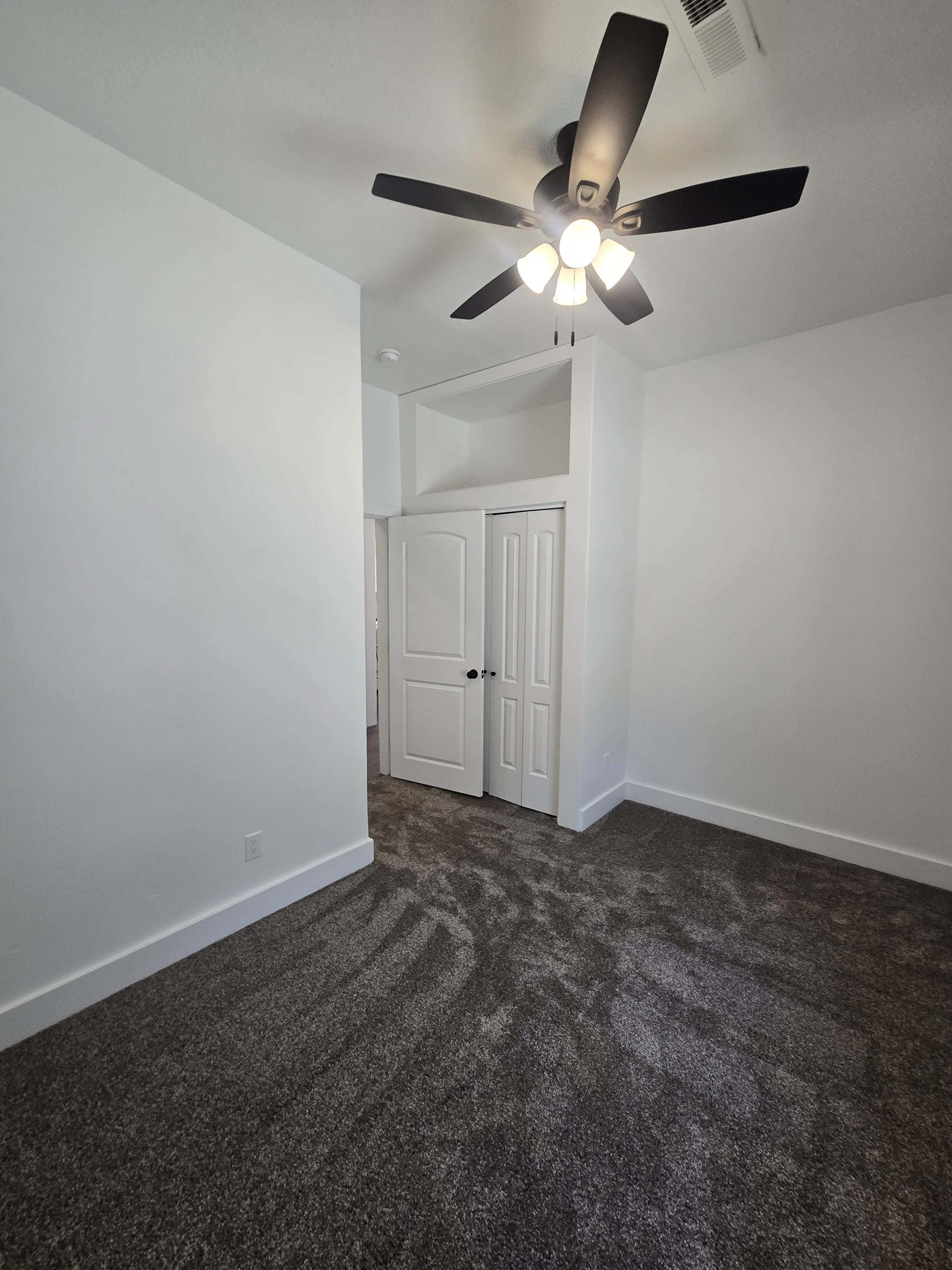Empty room with white walls, a ceiling fan with five black blades and light bulbs, beige carpet, and a partially open white closet door.
