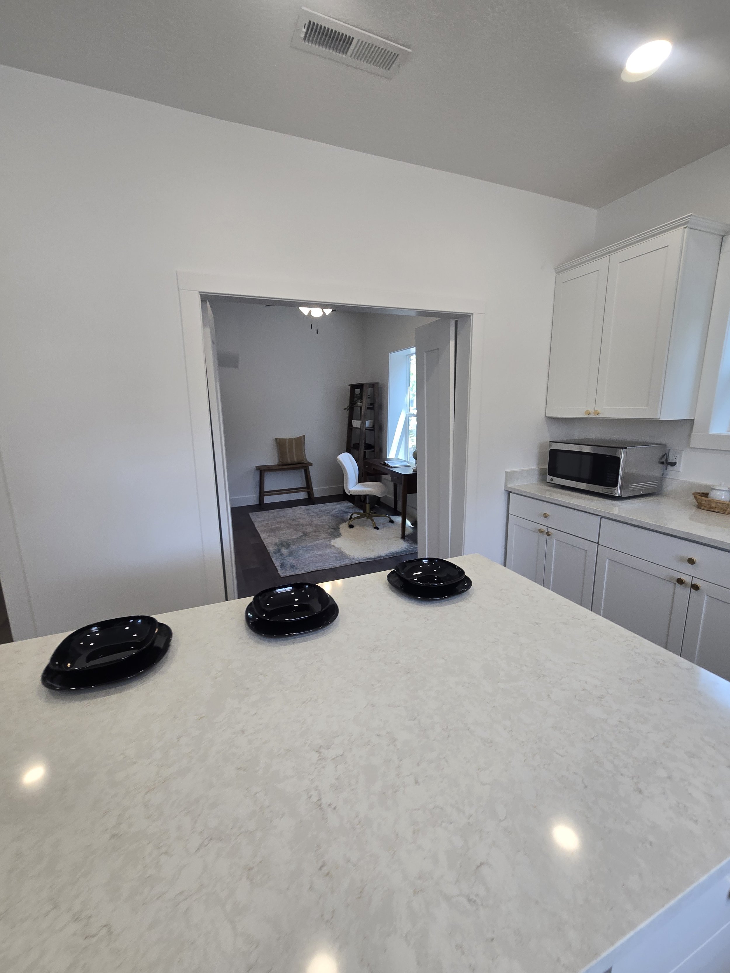 A white kitchen island with three black bowls and a black plate, a doorway leading to a room with a white office chair, a small table, a wooden shelf, and a window showing outside, white cabinets, and a microwave on the countertop.