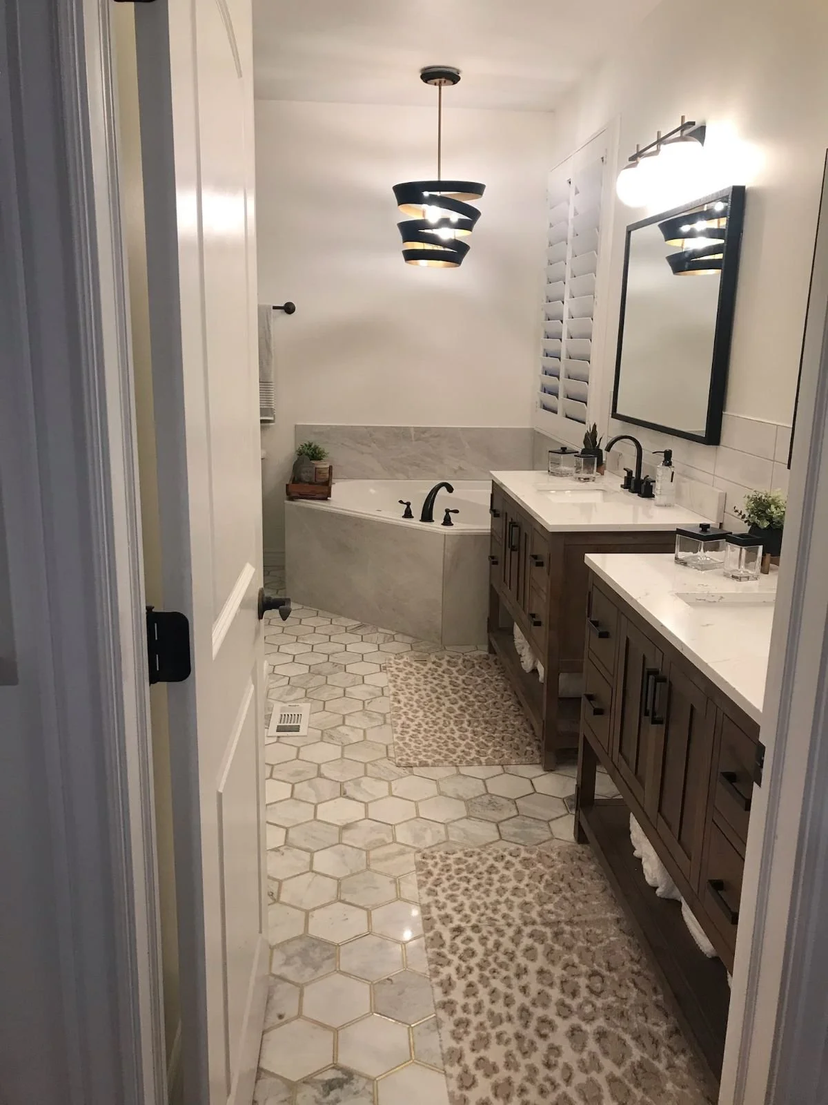 A bathroom with hexagonal tile flooring, a bathtub, double vanity with sinks and mirrors, and decorative rugs.