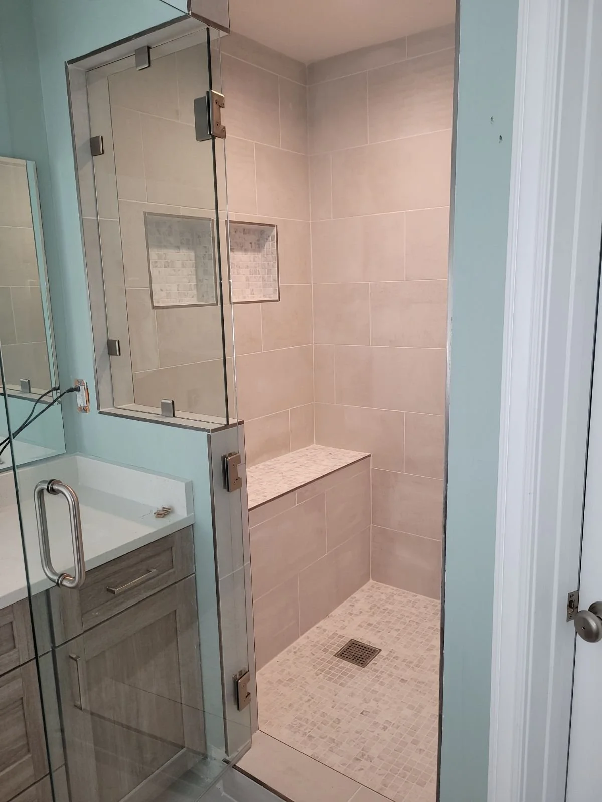 Bathroom shower with glass door, beige tiles, built-in bench, and mosaic tile floor, alongside a vanity with a mirror.
