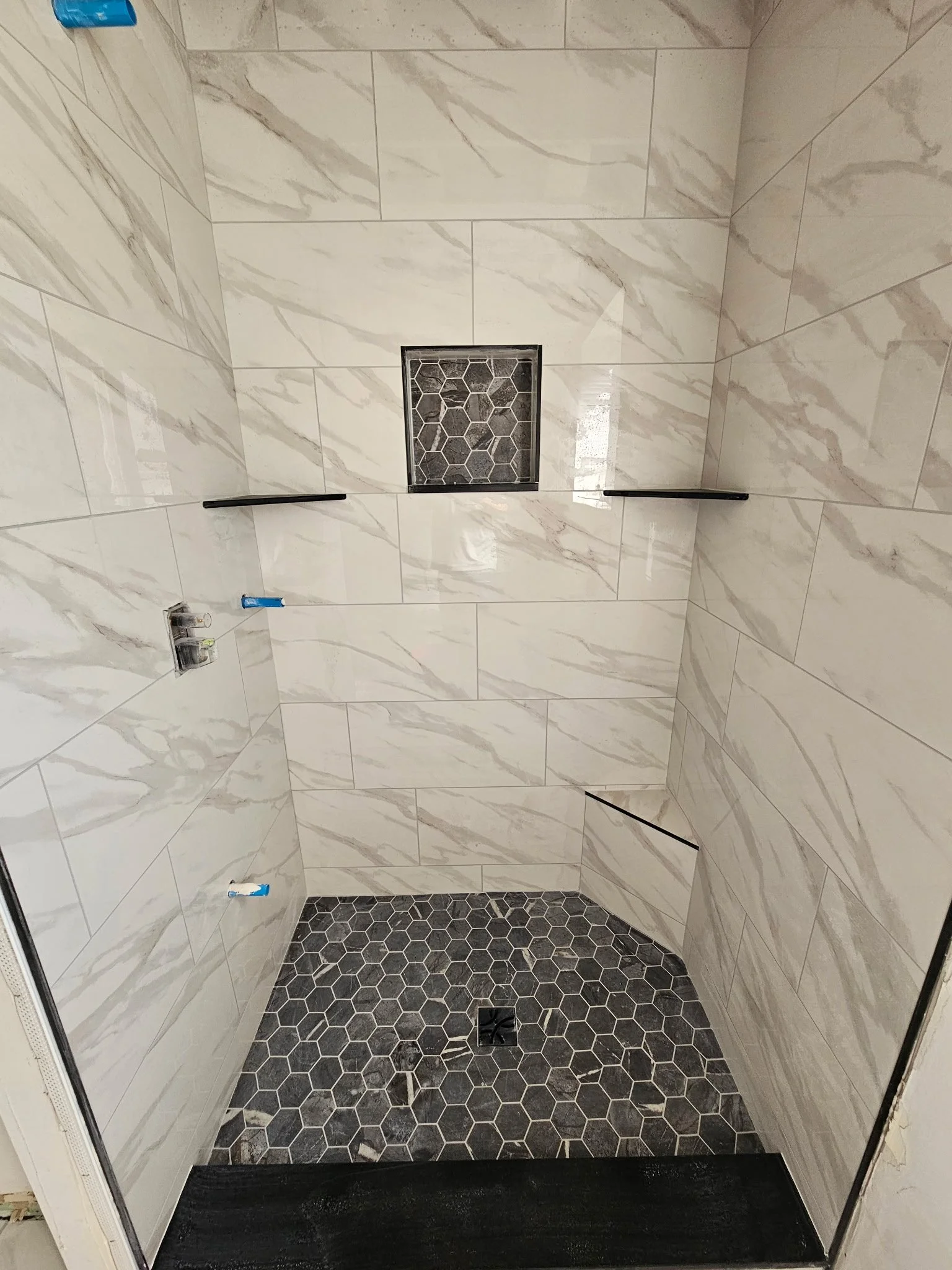 A newly renovated shower with large beige marble-like tiles on the walls and black hexagon floor tiles. It has a drain in the center, a square niche with hexagon tiles, and two black grab bars.