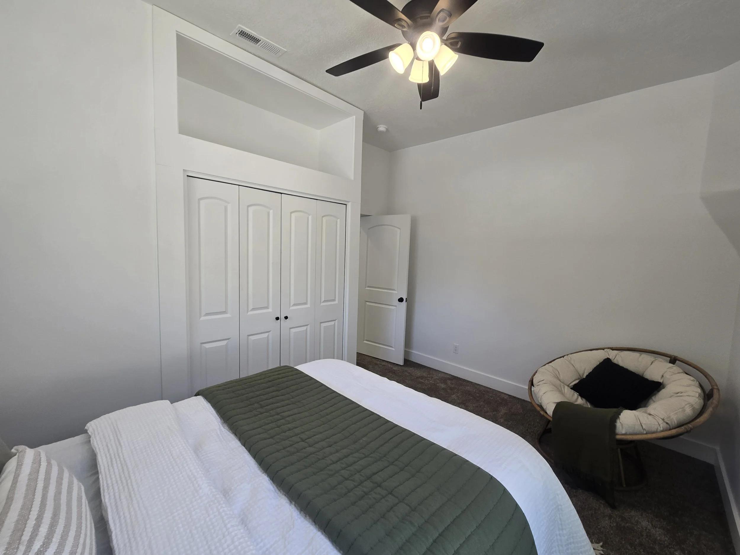 A bedroom with a bed, green and white bedding, a round papasan chair with a black pillow, white closet doors, white walls, a ceiling fan with lights, and a small balcony or loft area.