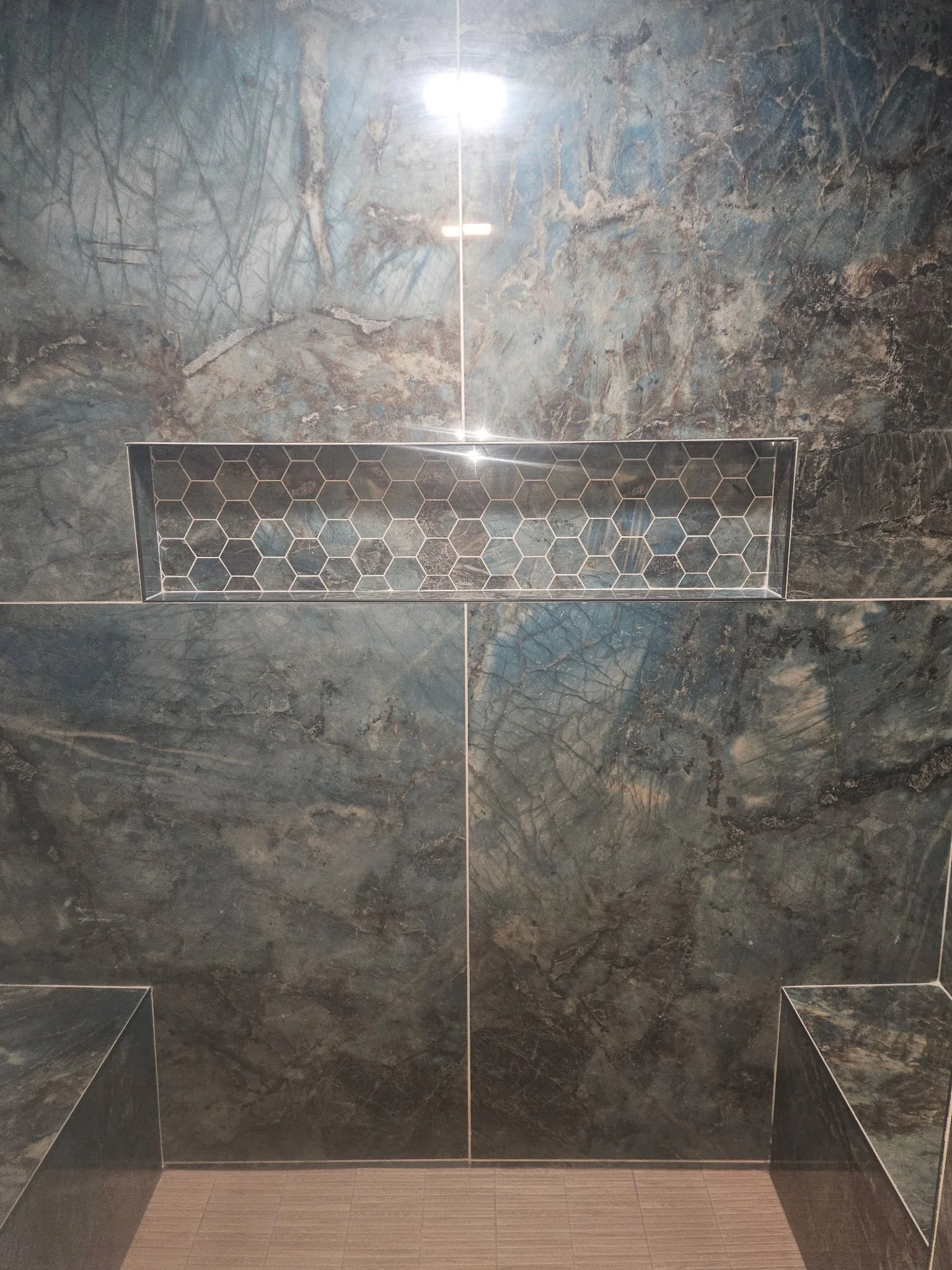 Reflection of ceiling lights on a polished dark marble wall with a rectangular metal vent with hexagonal pattern near the bottom.
