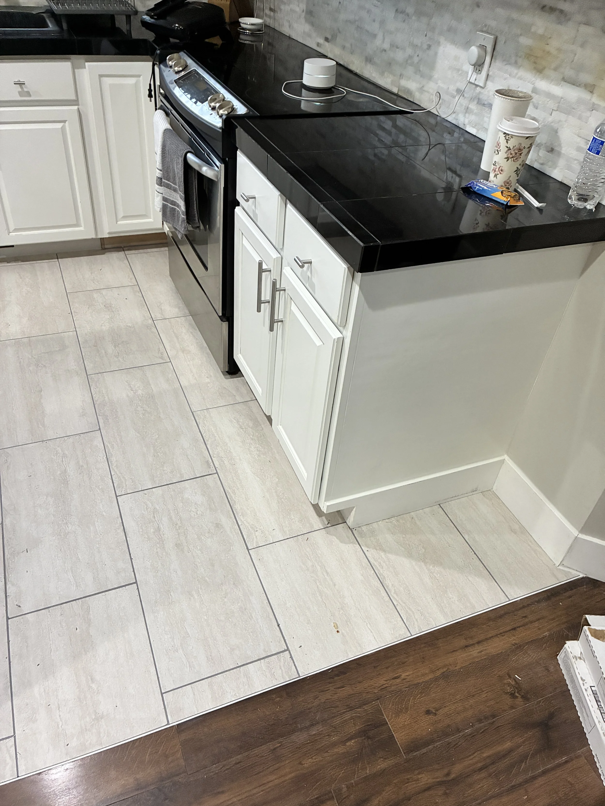 Kitchen floor with beige tiles transitioning to dark wood flooring, black countertop, and white cabinets. There is a black stove with a gray and white towel hanging on the oven handle. Various items are on the countertop, including a water bottle, a 