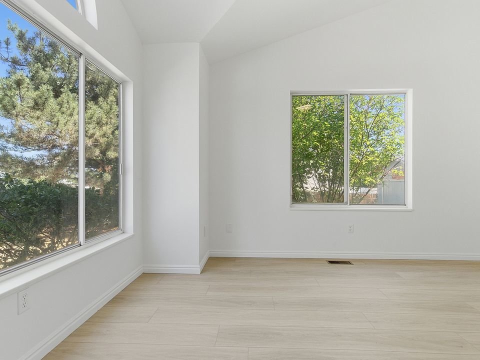 Empty room with white walls, light wooden floor, two large windows showing green trees outside, and a ceiling vent.