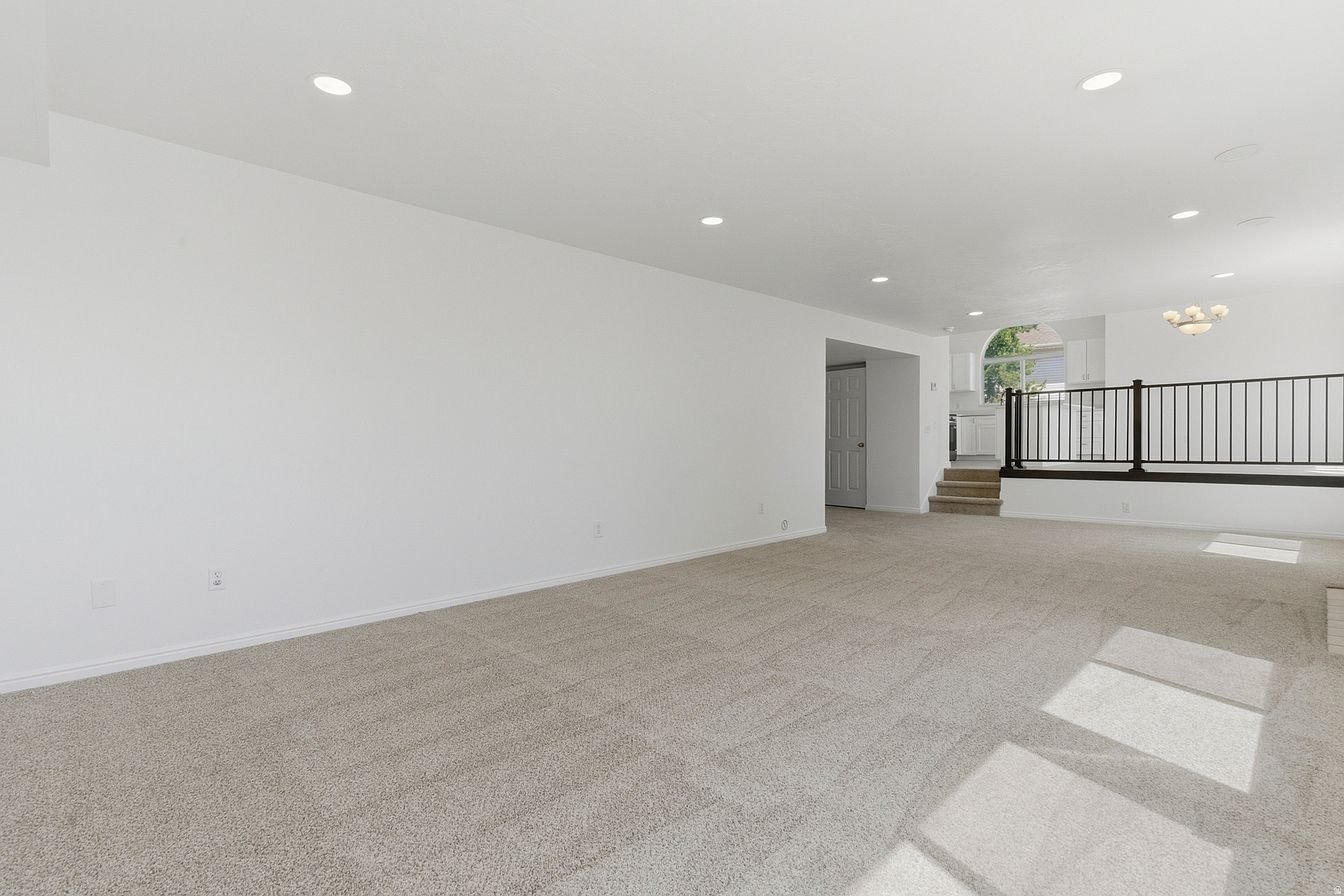 Empty living room with beige carpet, white walls, and ceiling, with overhead lighting and natural light from windows, leading to a kitchen area upstairs.
