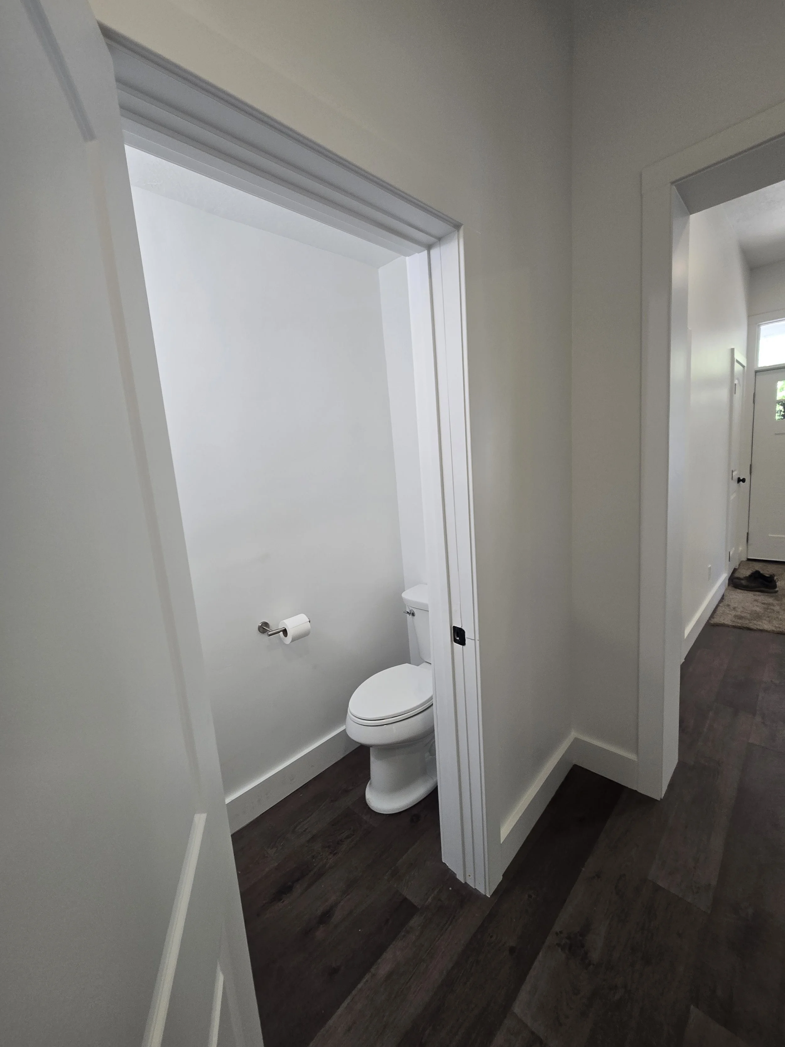 A small, partially open bathroom door reveals a white toilet inside a narrow space with white walls. The room has a toilet paper roll mounted on the wall. The floor is covered with dark wood-like flooring. Adjacent to the bathroom, a hallway with sim