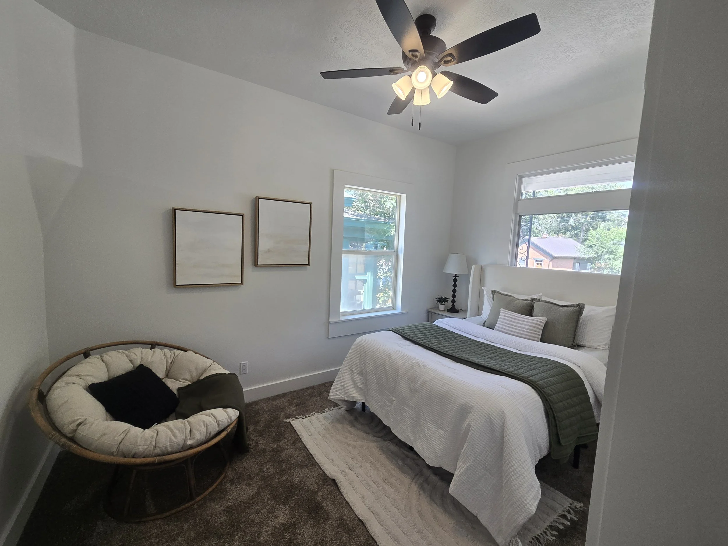 A bedroom with a white bed, green blanket, and decorative pillows, a standing lamp, two windows with views of trees and a neighboring house, two framed art pieces on the wall, a ceiling fan with light, a small round chair with cushions, a striped rug