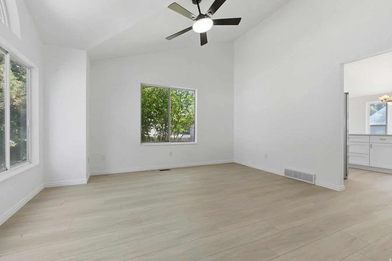 Empty room with white walls, light wood flooring, windows on two sides, a ceiling fan with lights, and an opening leading to a kitchen area.