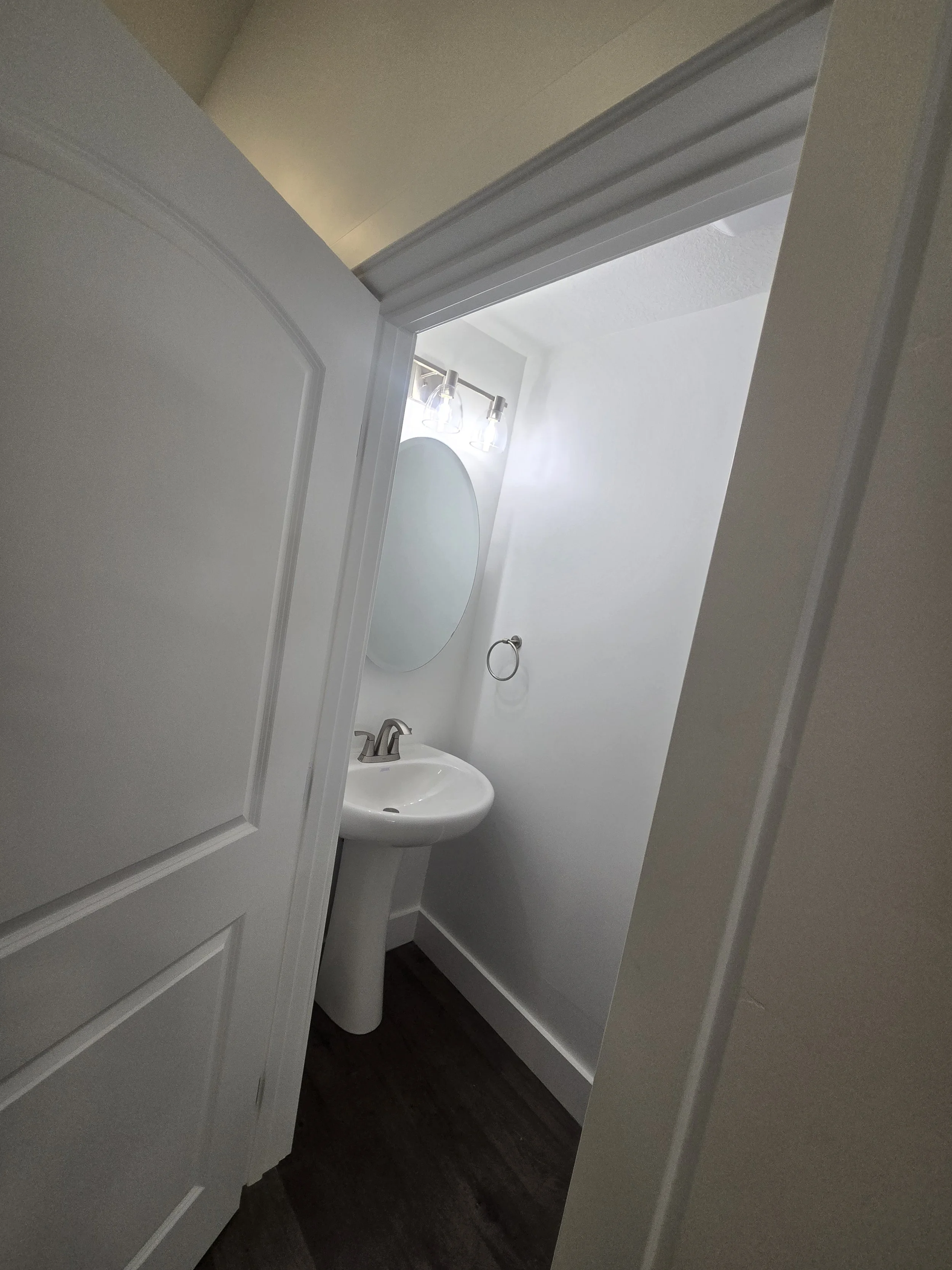 A small bathroom with a pedestal sink, oval mirror, and two light fixtures above the mirror, visible through an open door.