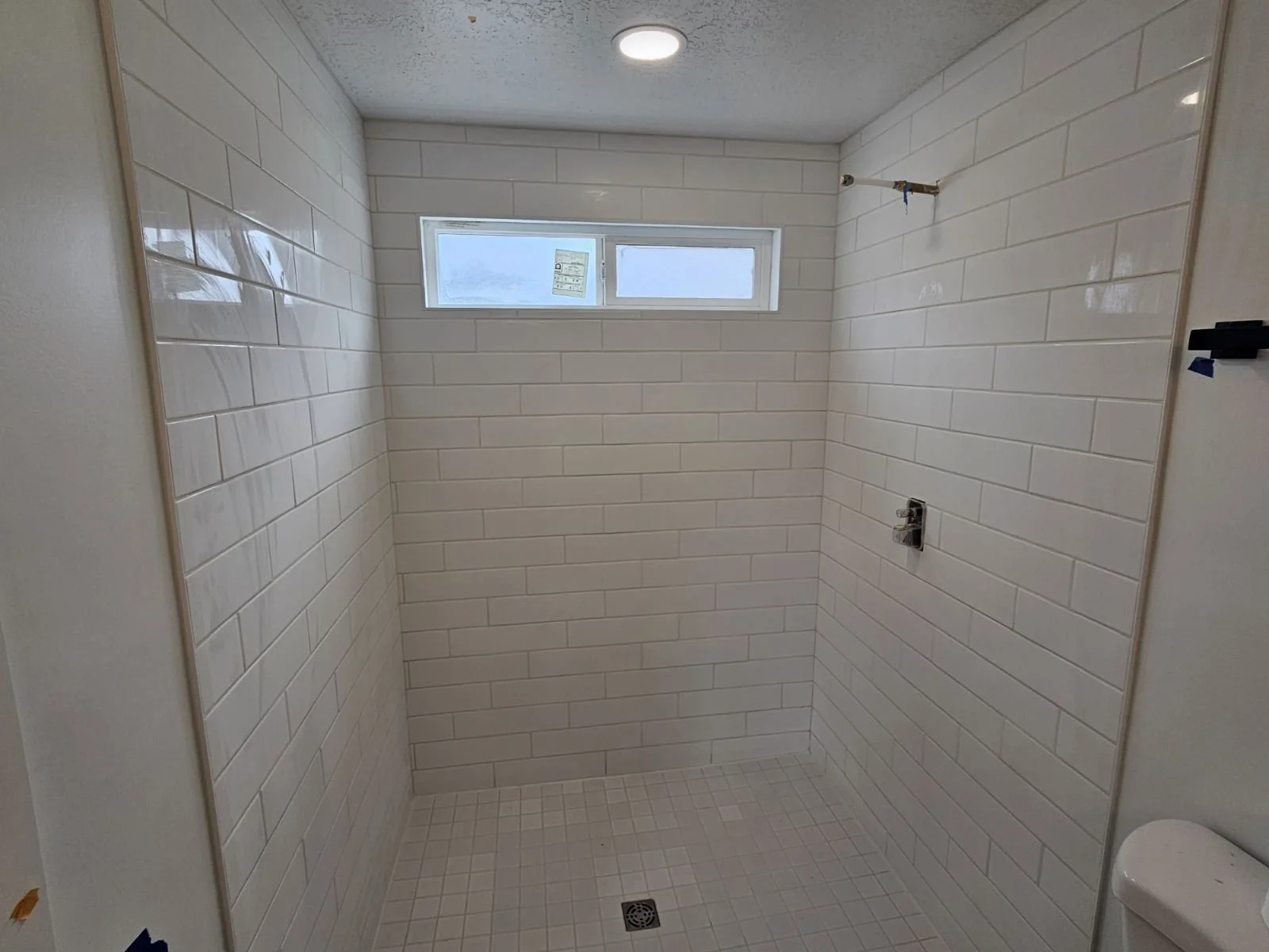 Unfinished shower with white subway tile walls, small square floor tiles, a small window, and a ceiling light.