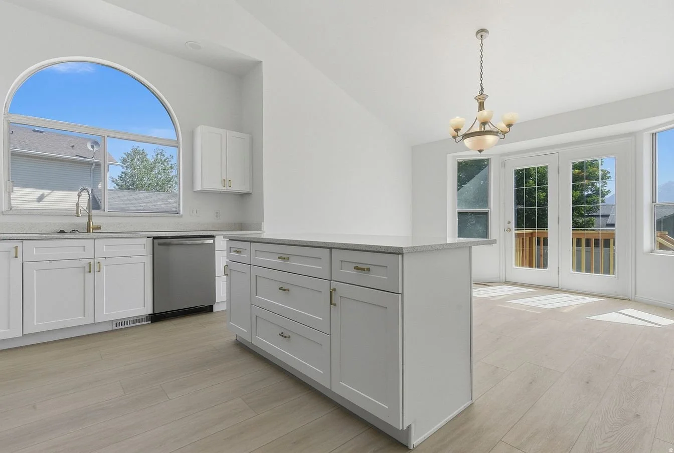 Bright kitchen with white cabinets, large arched window, small island, and double glass doors leading to outside deck with trees