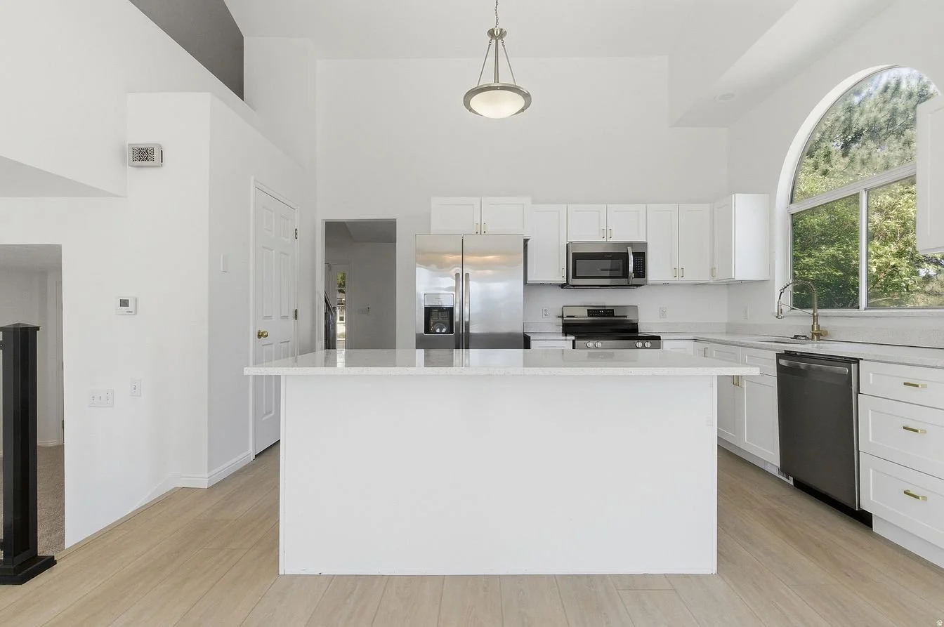 Empty white kitchen with stainless steel appliances, black dishwasher, and large arched window showing outdoor trees.
