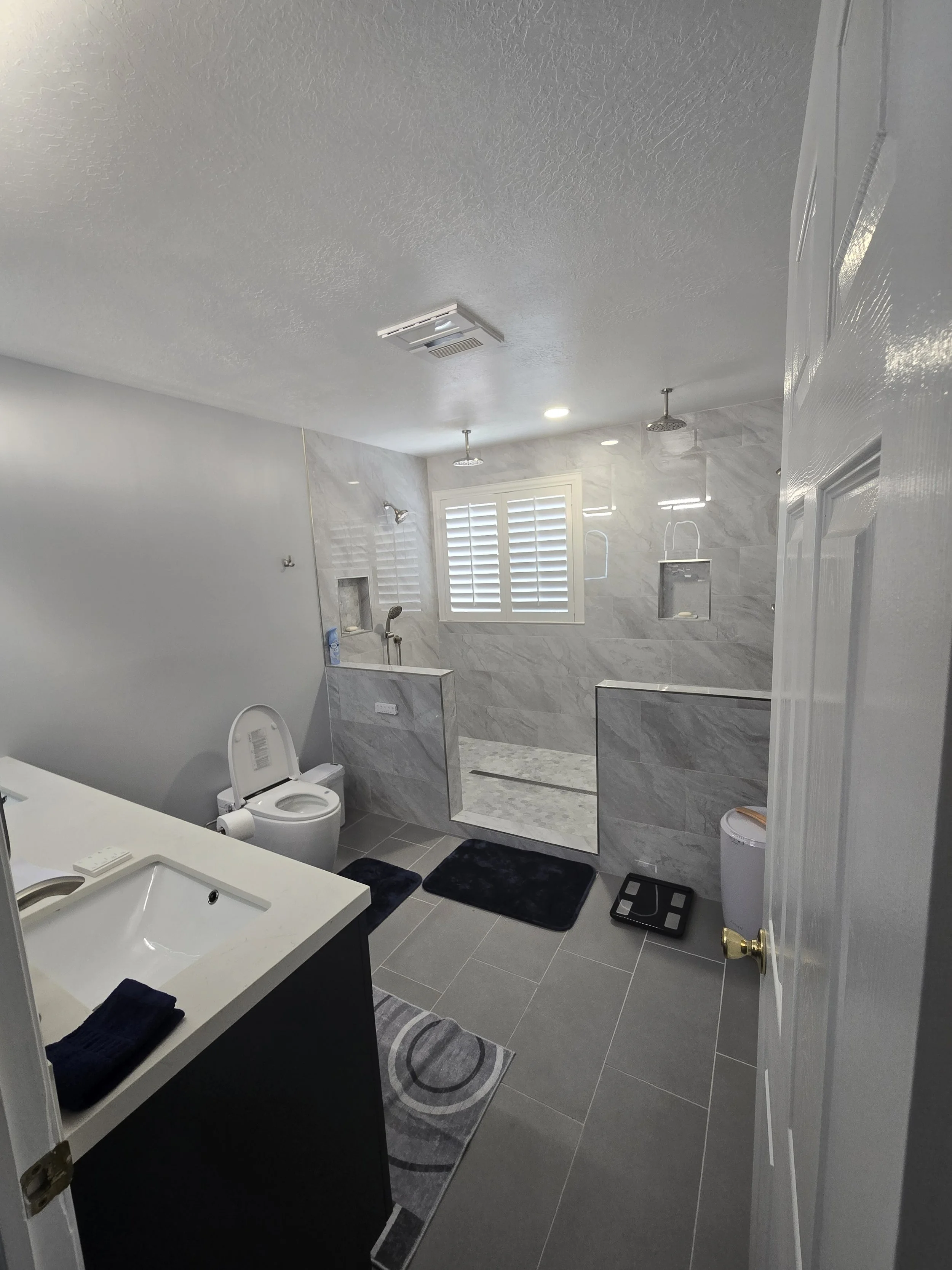 A modern bathroom with gray tiled flooring and walls, a white toilet, a vanity with dark cabinet and white countertop, a rectangular mirror, black towels, black slippers, and a gray and black rug. The shower area features marble tiles, two showerhead
