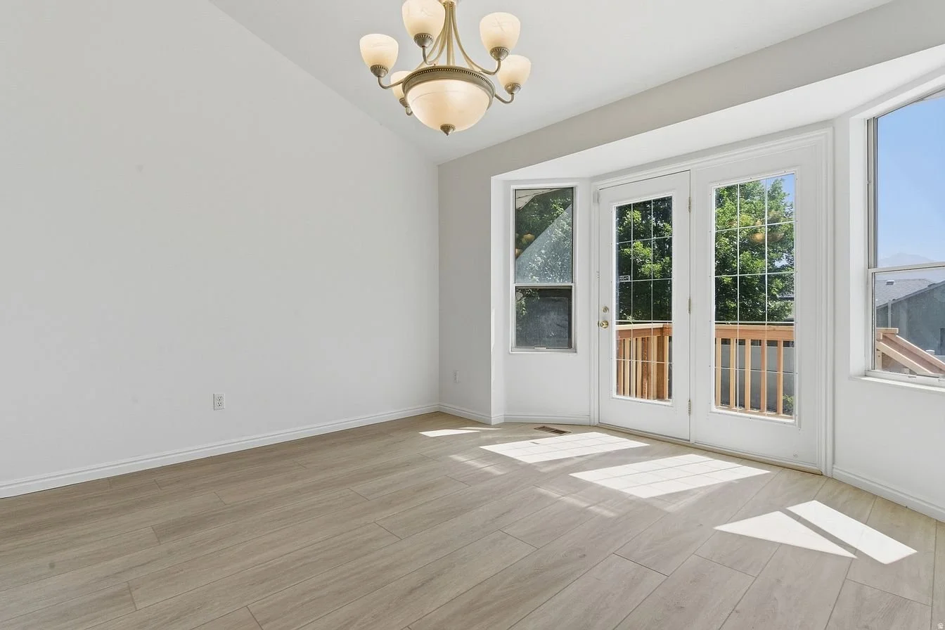 Empty bright room with large windows, a chandelier, and light wooden floors.
