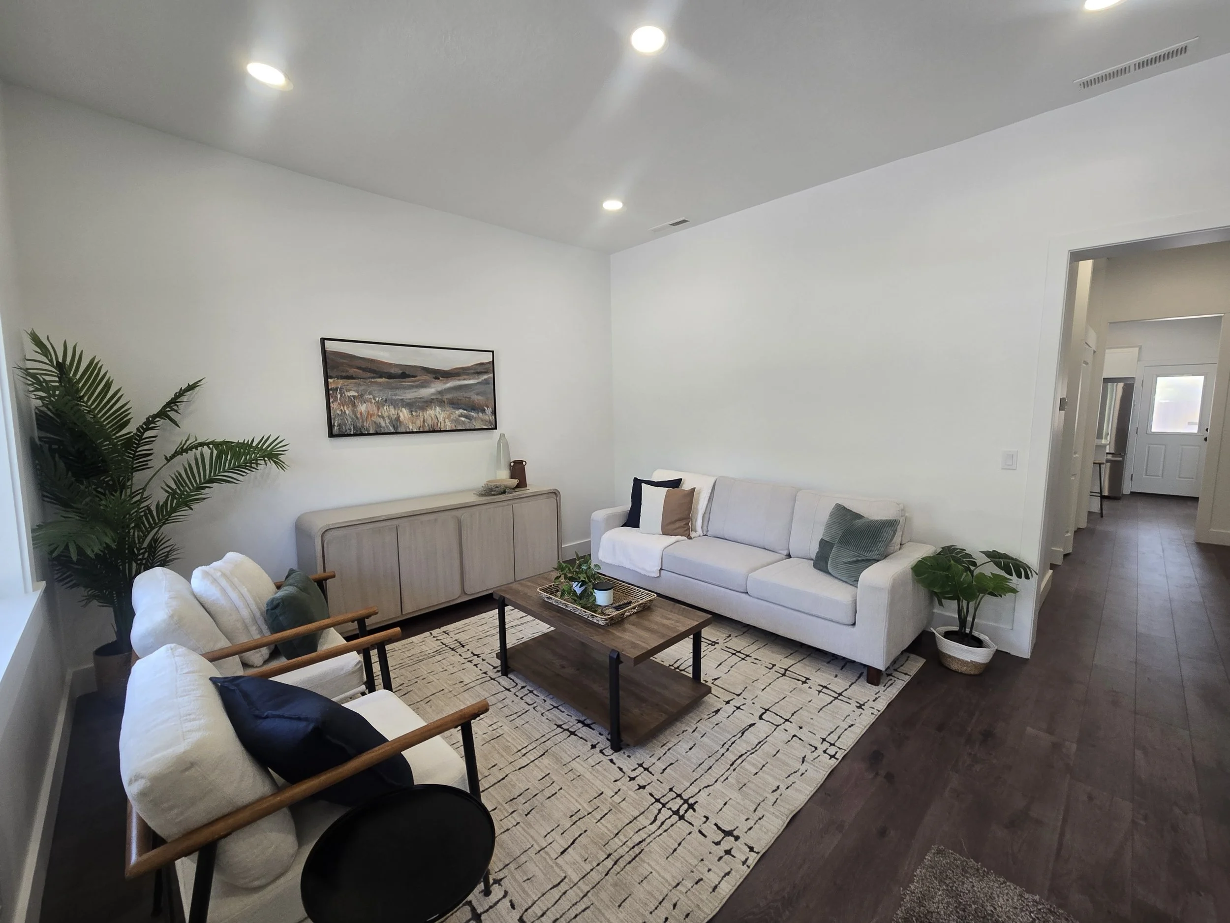 Living room with white sofa, patterned area rug, wooden coffee table, potted plants, wall art, and chairs along the wall.