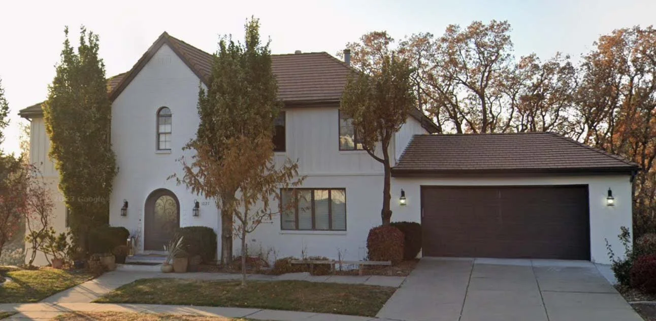 A two-story house with white exterior walls, dark roof, and attached garage, surrounded by trees and front yard.