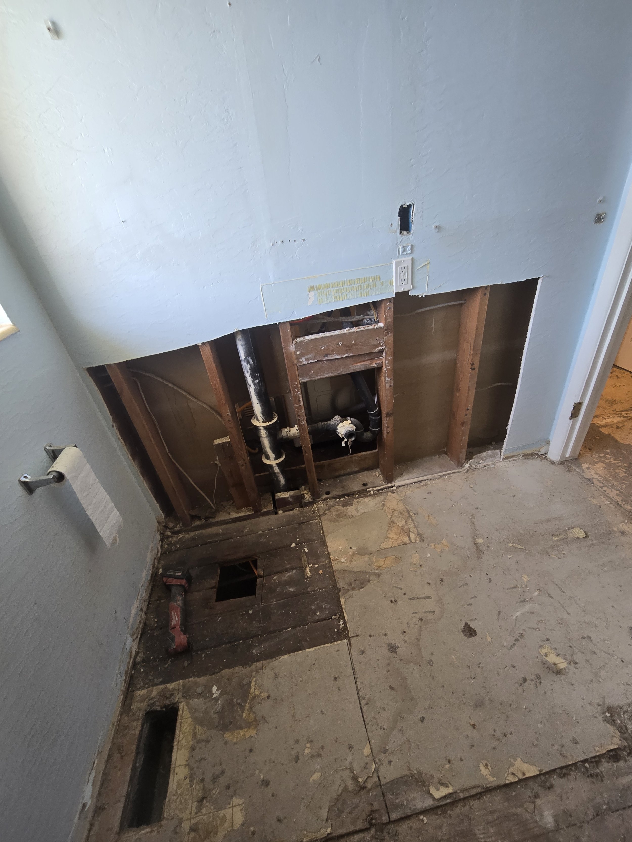 A partially demolished wall showing exposed wooden studs and plumbing pipes, with drywalled sections and an electrical outlet visible.