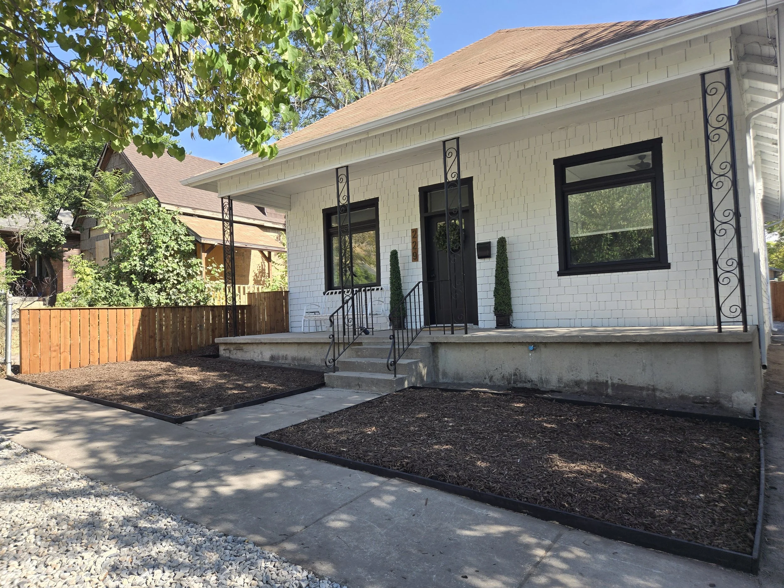 A white house with black window frames and a black front door, front porch with decorative black metal columns, surrounded by green trees and a fenced yard, with a clear blue sky overhead.
