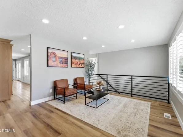 Living area with two brown chairs, black coffee table, framed landscape artwork, potted plant, large window with white blinds, and a black metal railing in a modern home.