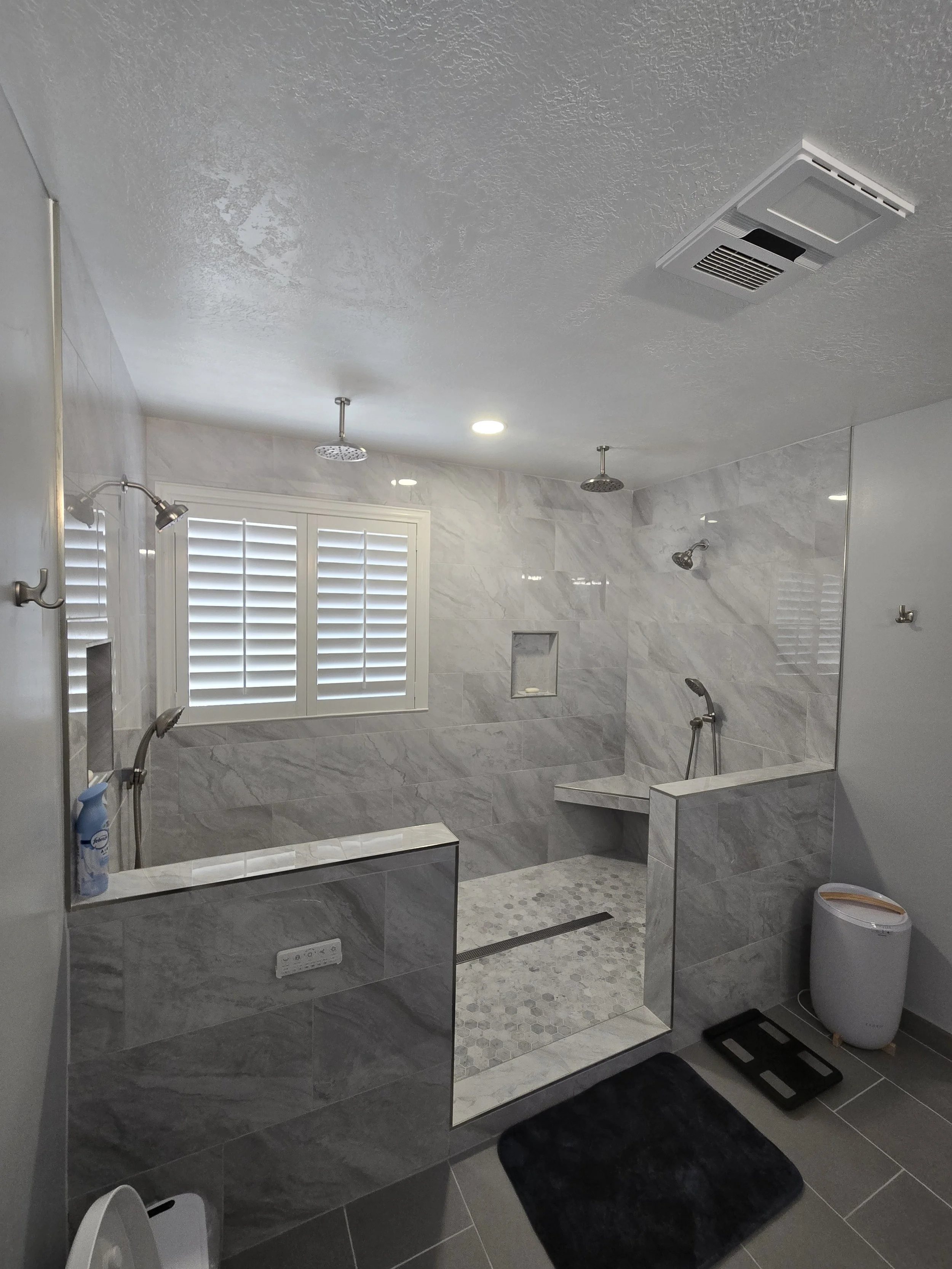 Bathroom with a walk-in shower featuring gray marble tiles, a bench, and multiple showerheads. Contains a window with white shutters, a white trash can, a black scale, and a towel hook.